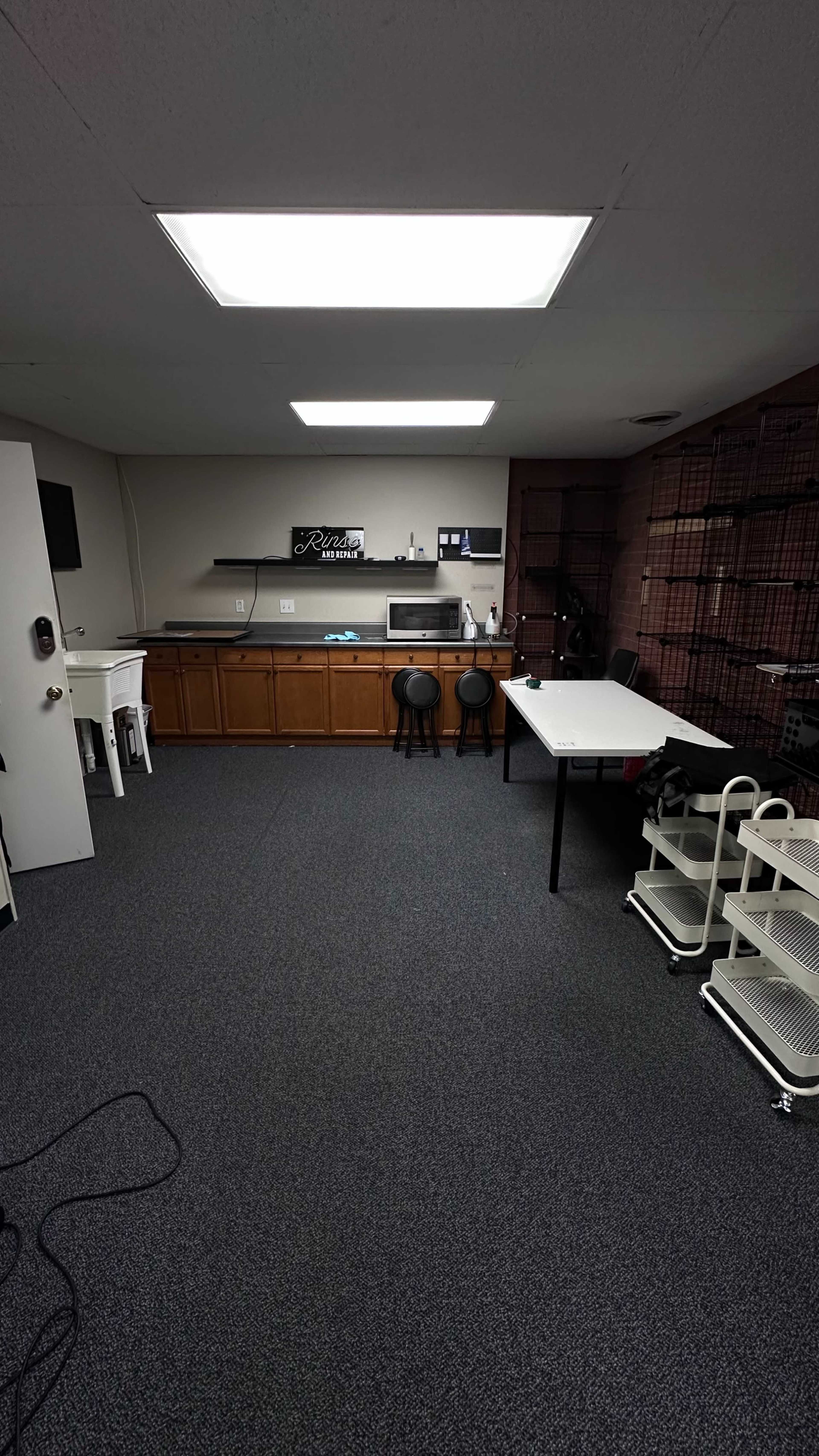The image shows a spacious room with a kitchenette area, a white table, black stools, and a storage unit with wheels.