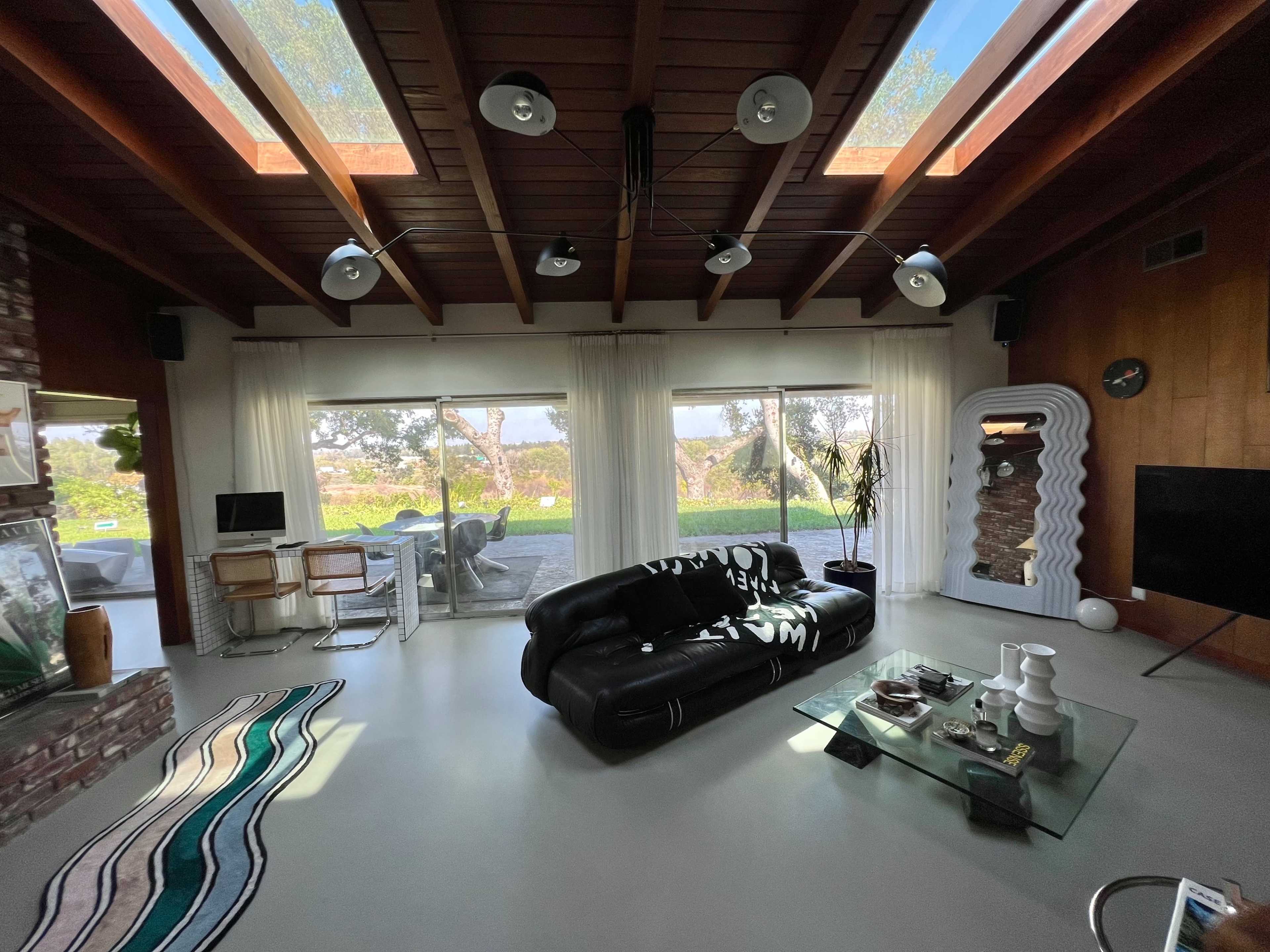 The image shows a modern living room with a black leather sofa, large windows allowing natural light, and a glass coffee table.
