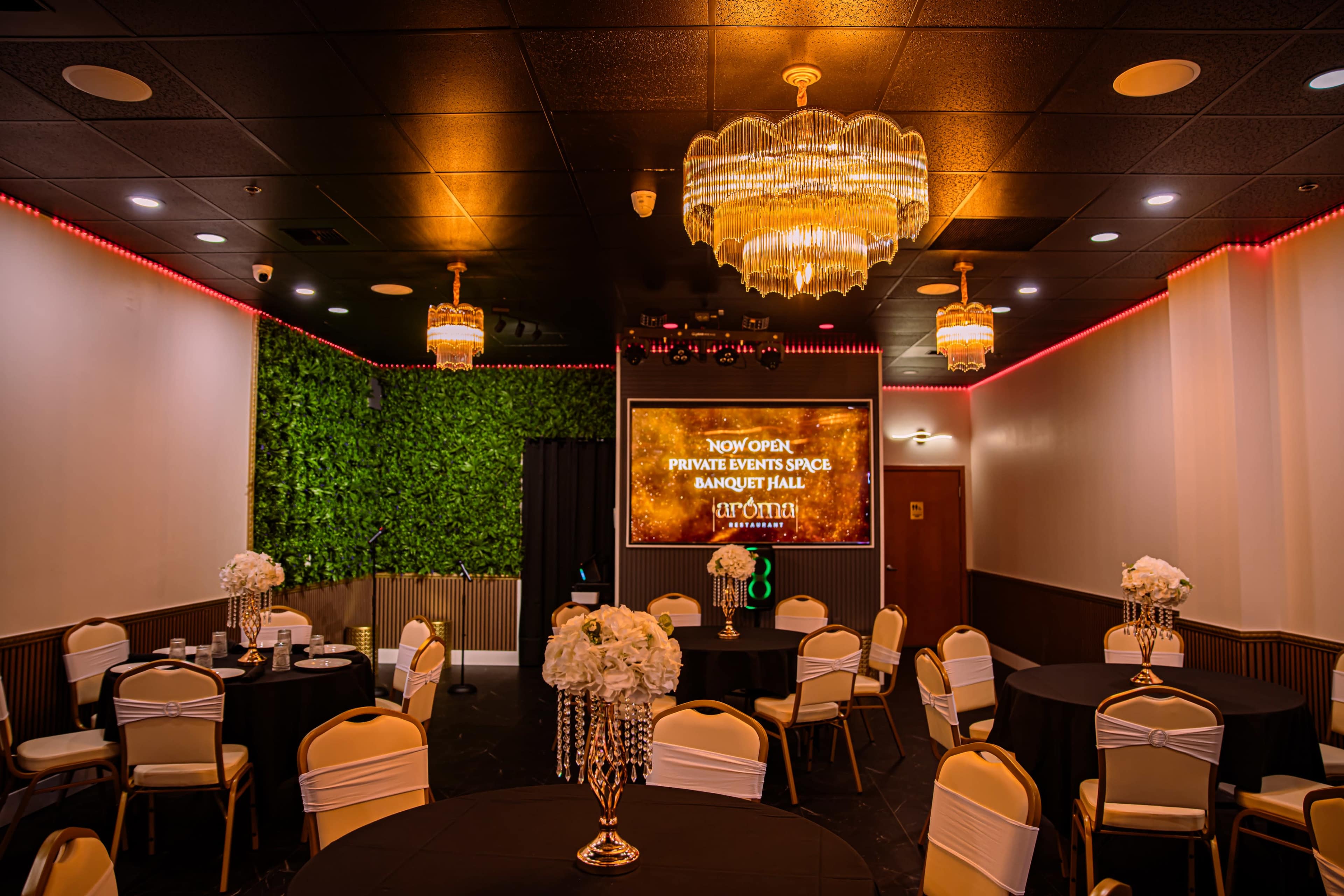The image shows a banquet hall with round tables covered in black cloth, adorned with white floral centerpieces, and features a large screen displaying "NOW OPEN: PRIVATE EVENTS SPACE" against a backdrop of greenery and elegant chandeliers.