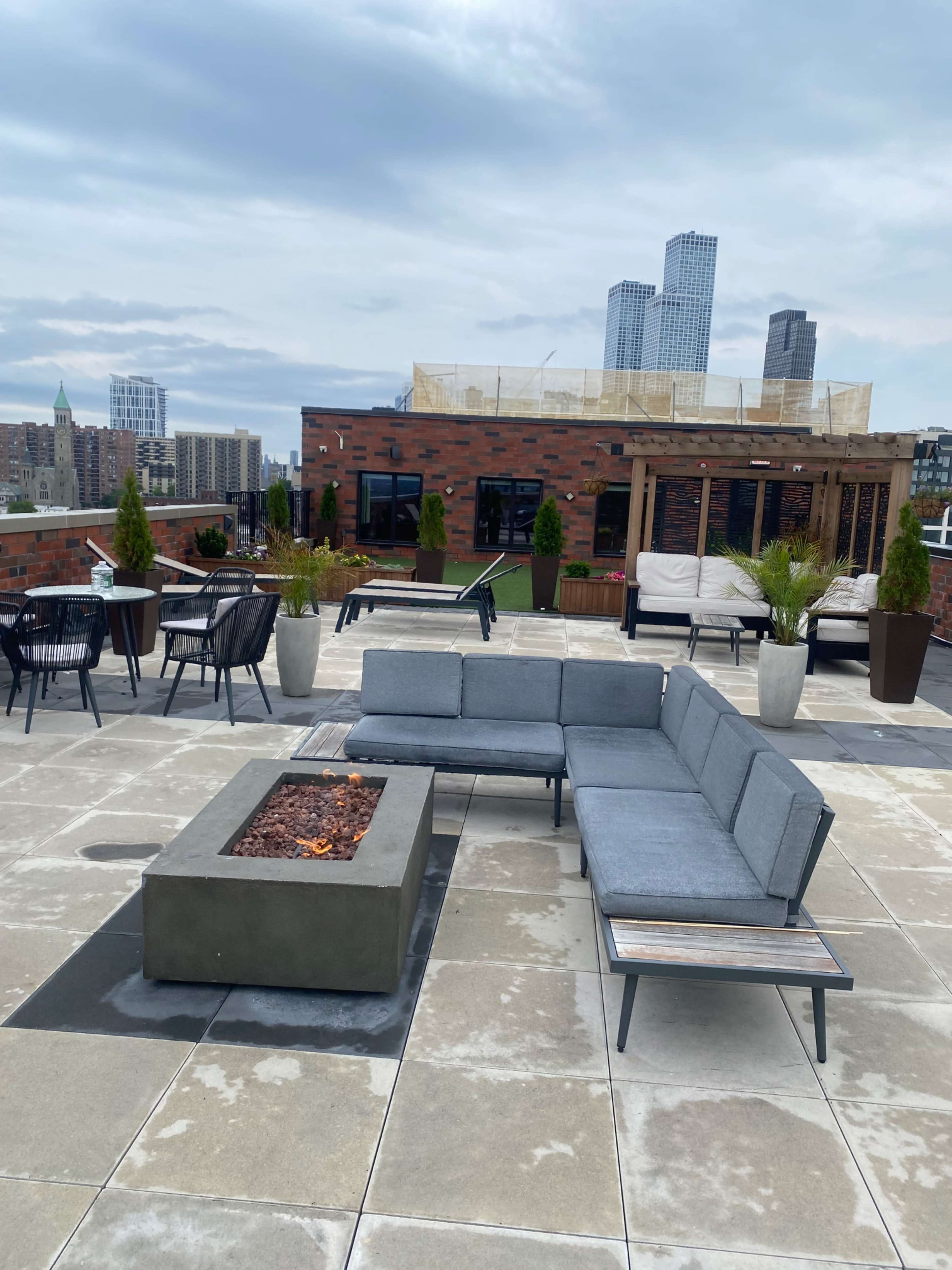 A rooftop terrace features a sectional sofa surrounding a fire pit, with city skyscrapers in the background.