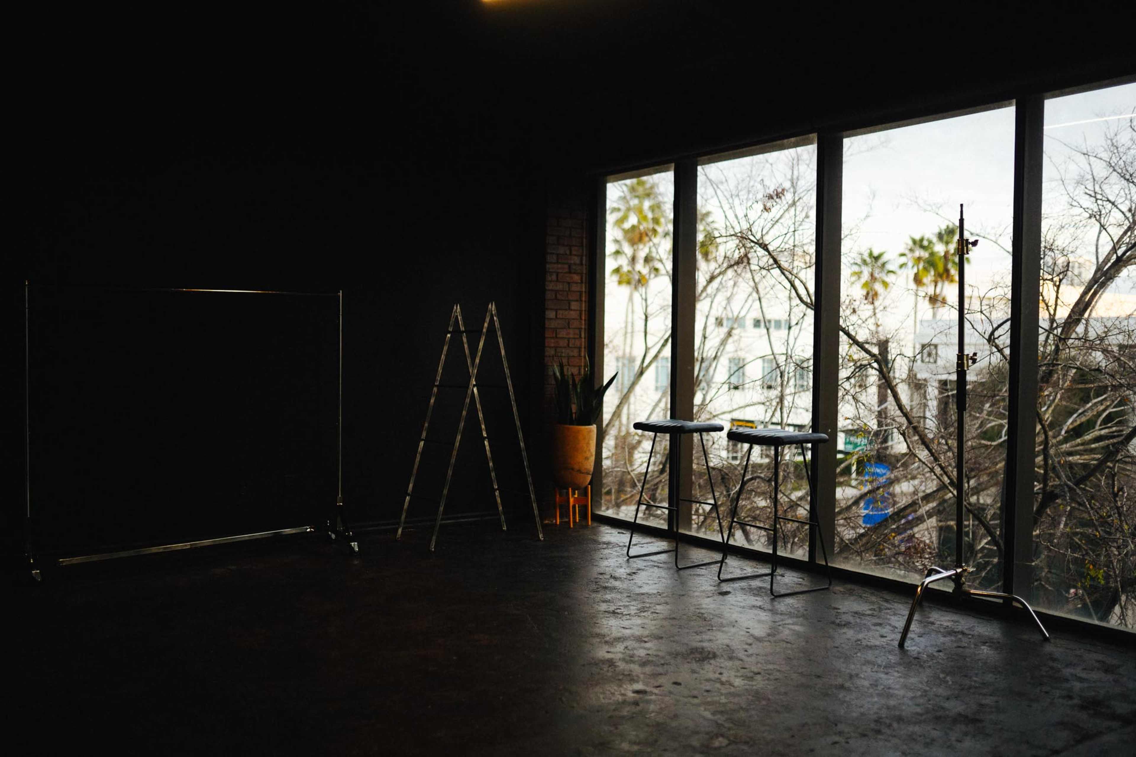 The image shows an empty room with black walls, a clothing rack, a ladder, two tables, and large windows revealing palm trees outside.