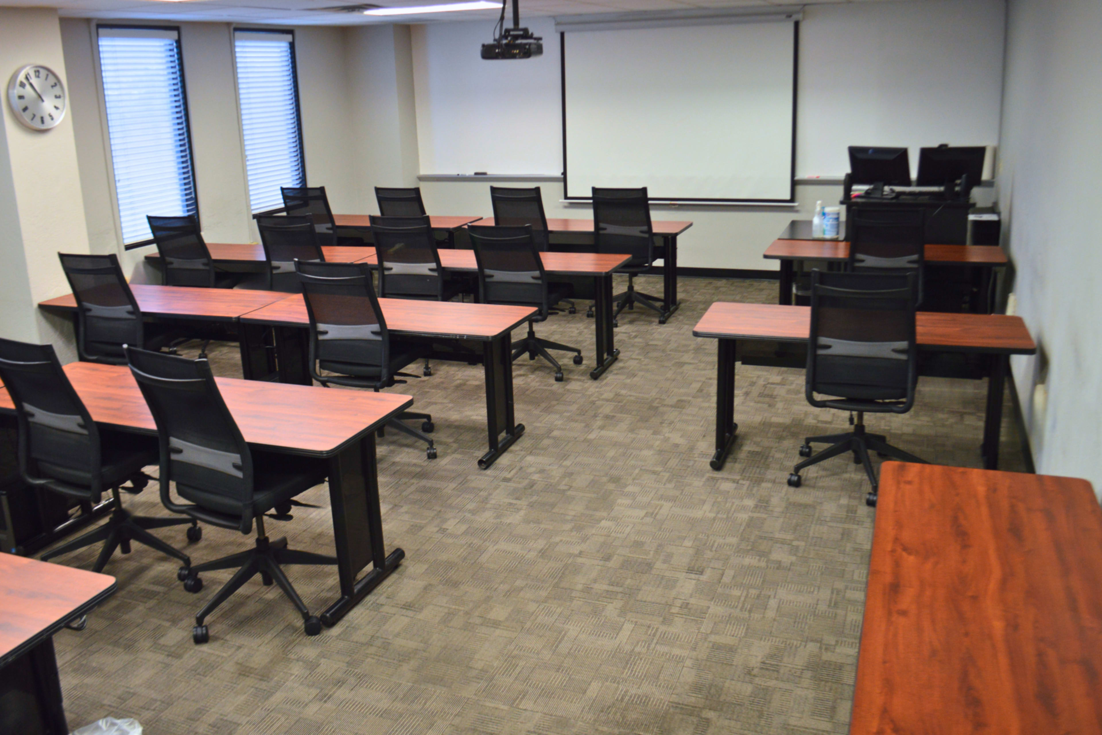 A classroom with several rows of desks and chairs arranged for seating, featuring a whiteboard and projector screen at the front.