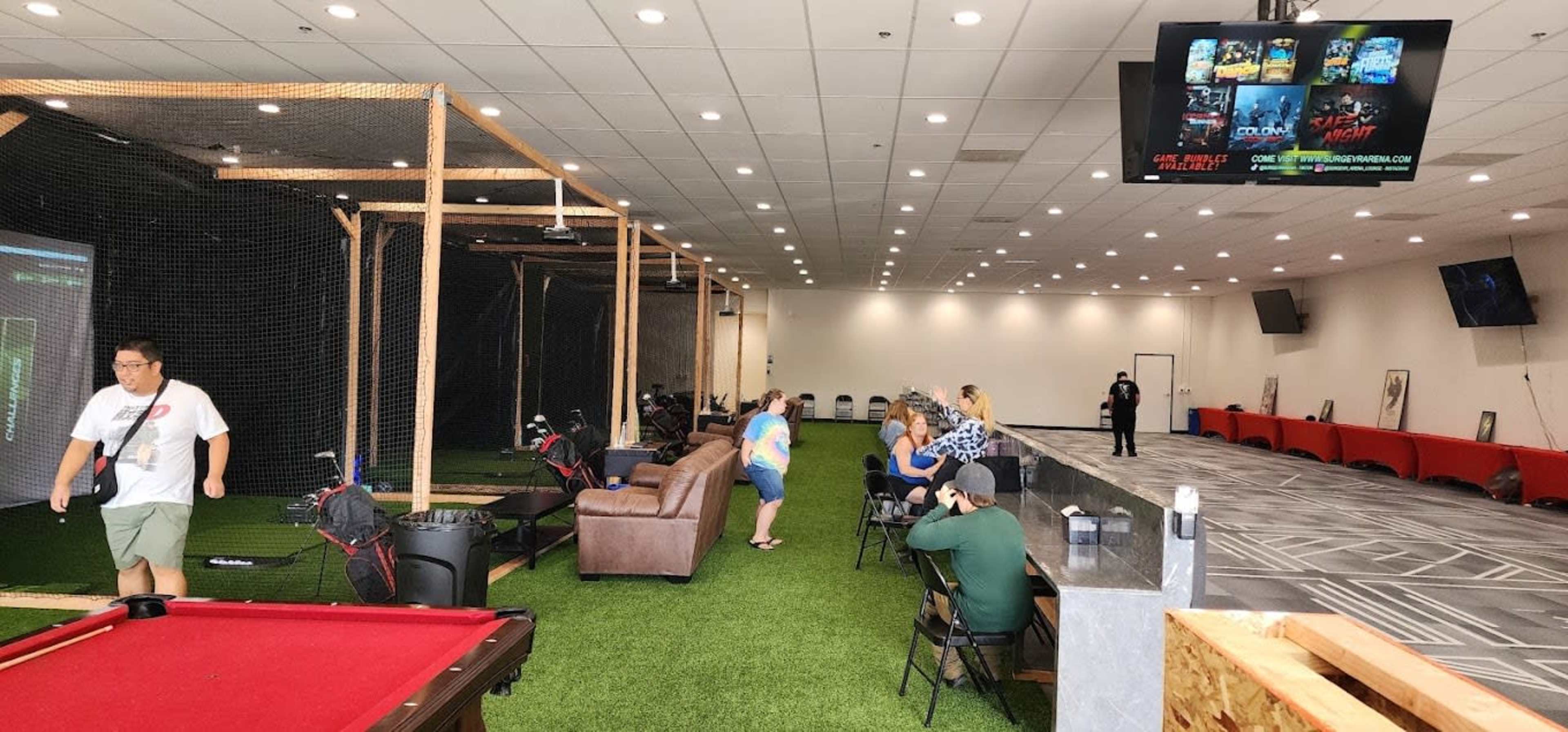 The image shows an indoor recreational space featuring a golf simulator area on the left, a pool table, seating for patrons, and a large screen displaying various games.