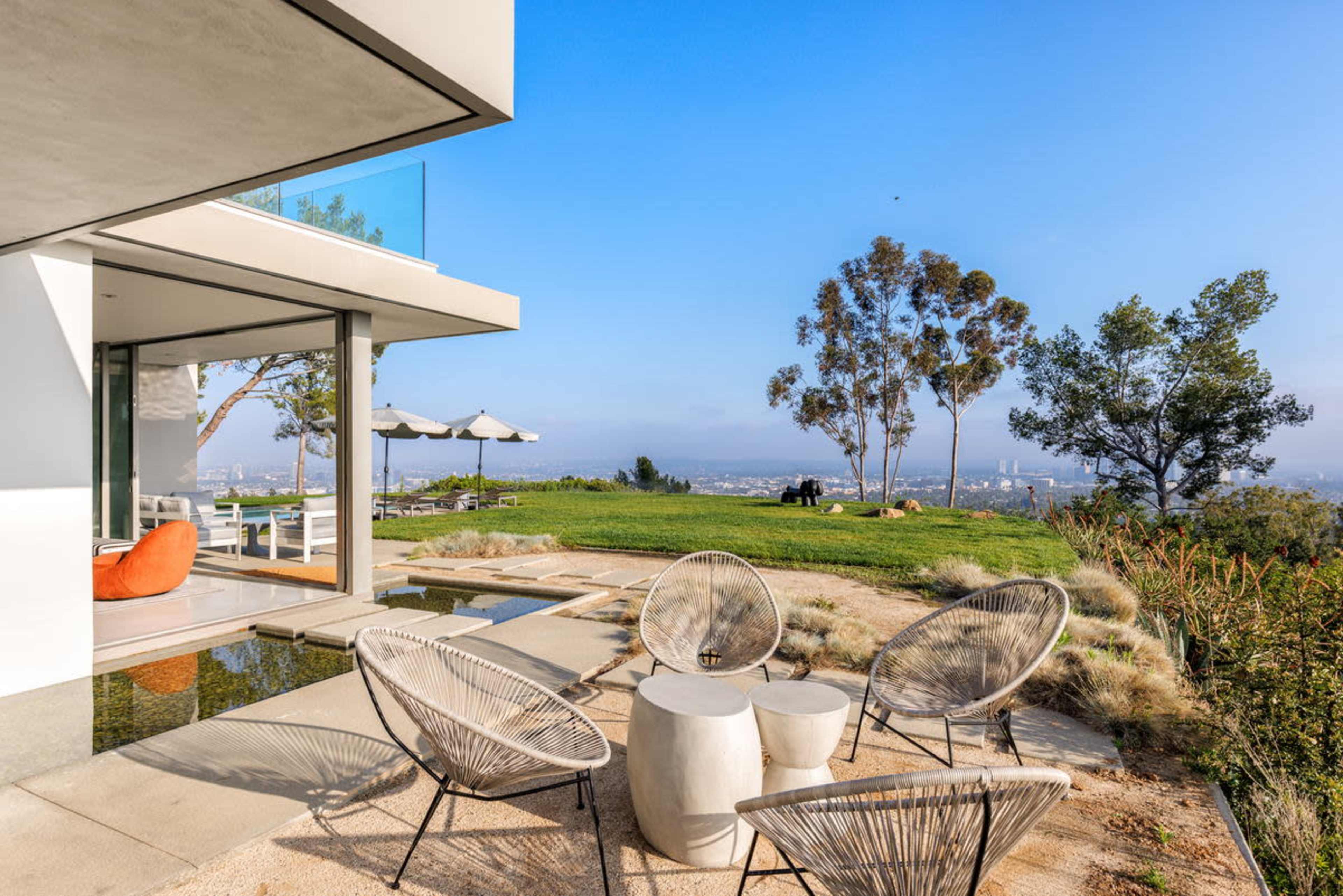 A modern outdoor seating area with circular stone tables and wire chairs overlooks a landscaped yard and city view.