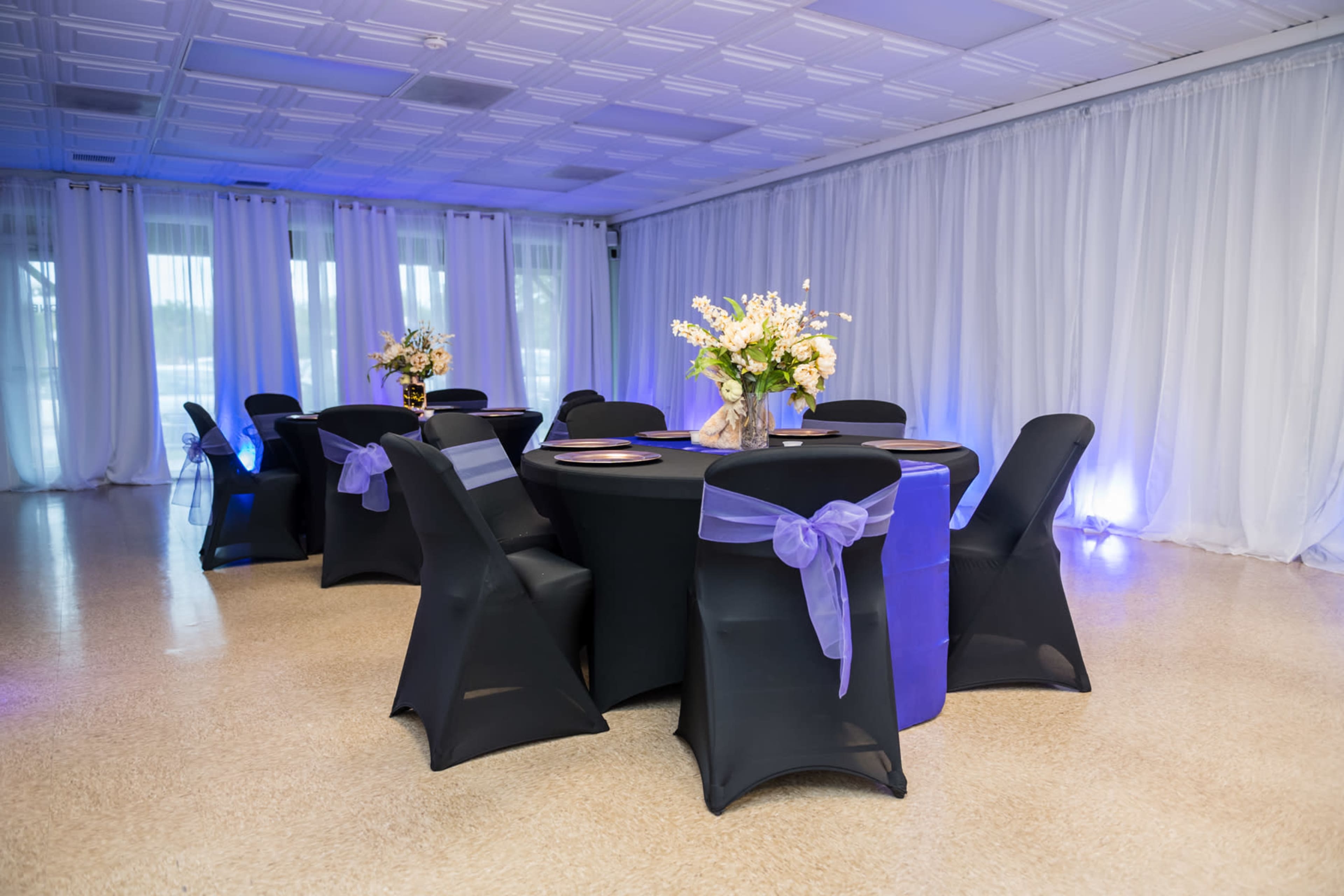 A banquet room features round tables with black tablecloths and lavender sashes, adorned with floral centerpieces, against a backdrop of white drapes.