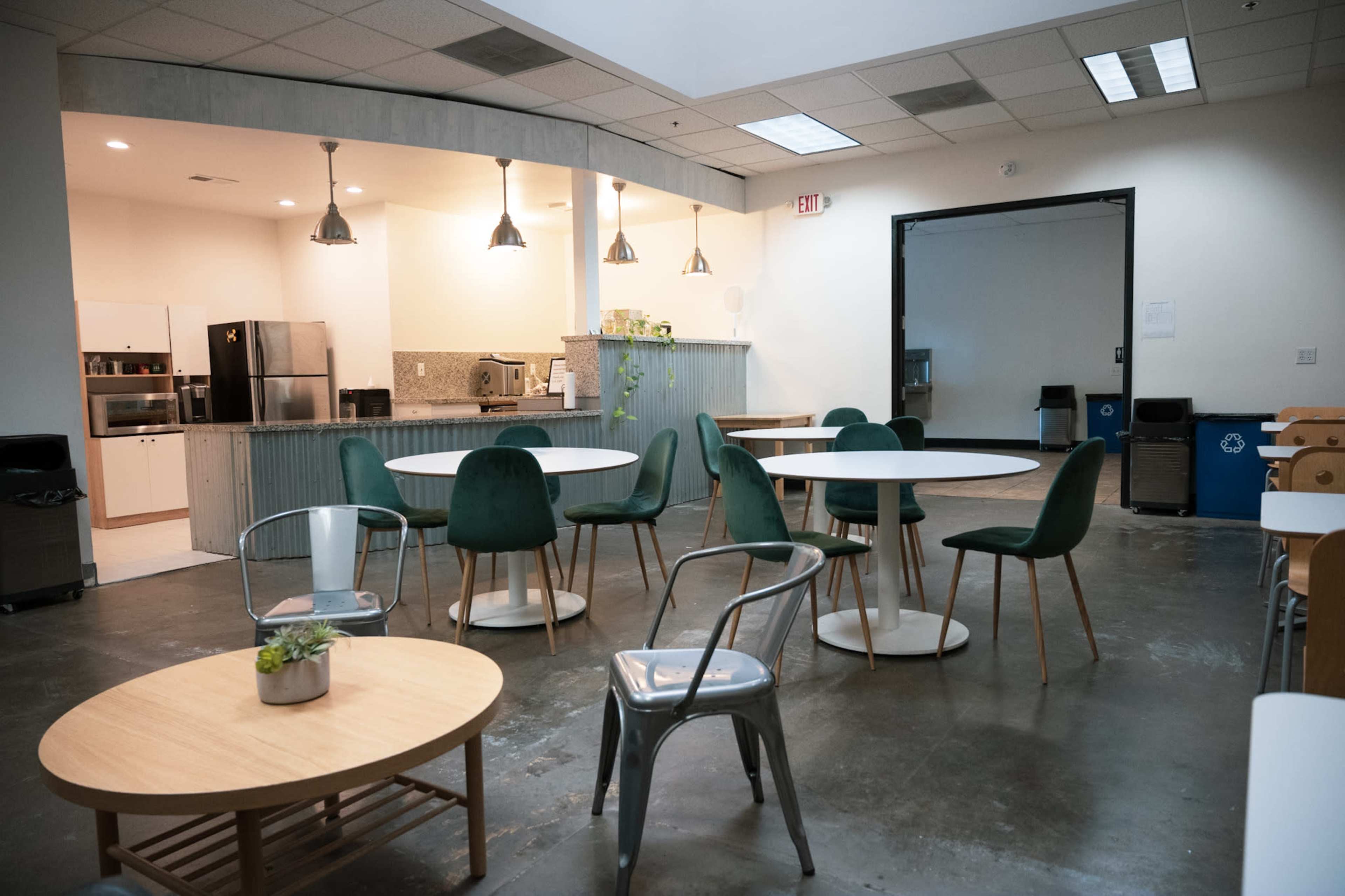 A modern break room with several tables and chairs, a kitchenette in the background, and various kitchen appliances visible.