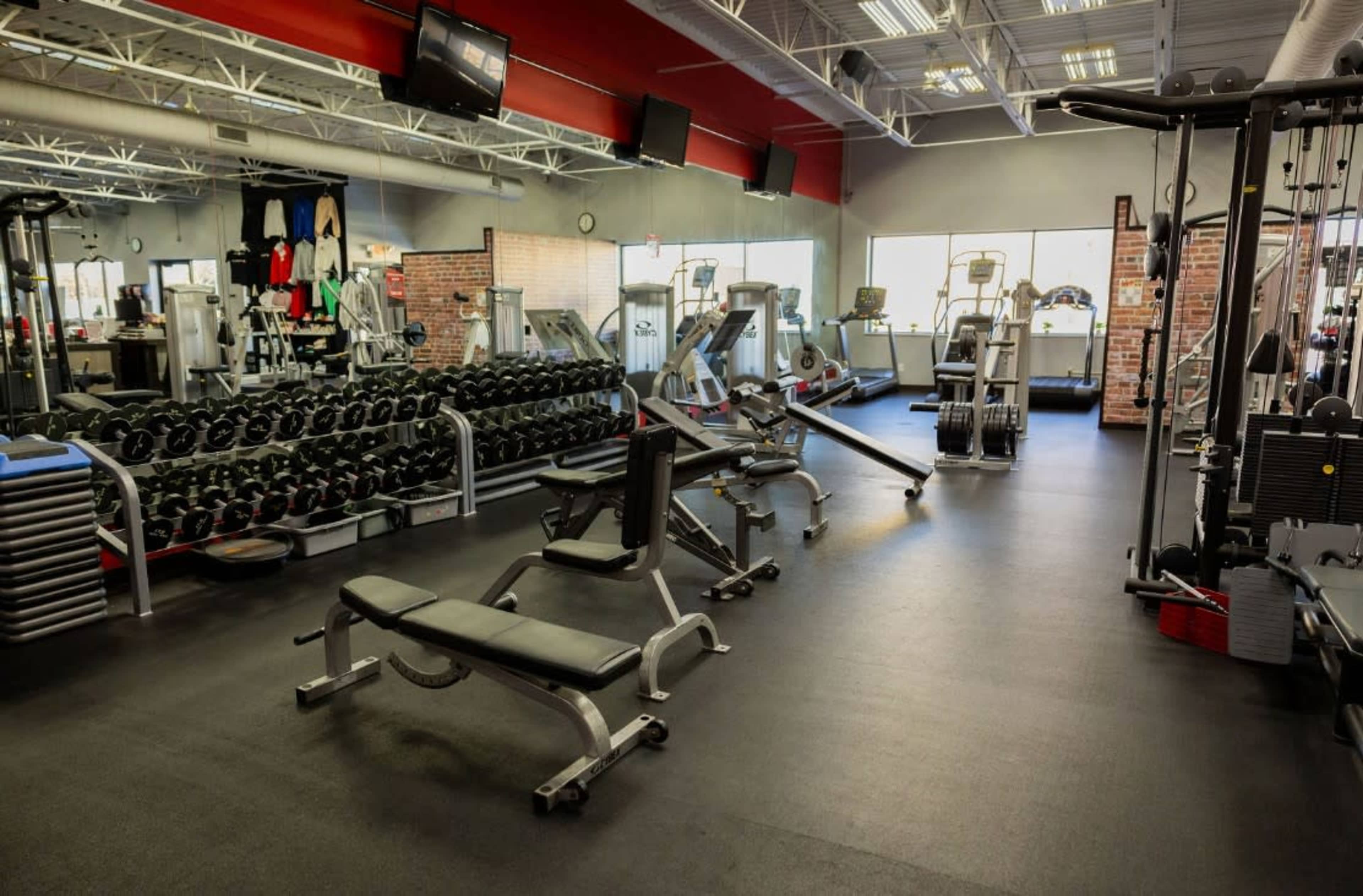 The image shows an interior view of a gym with various weightlifting equipment, cardio machines, and a row of dumbbells.