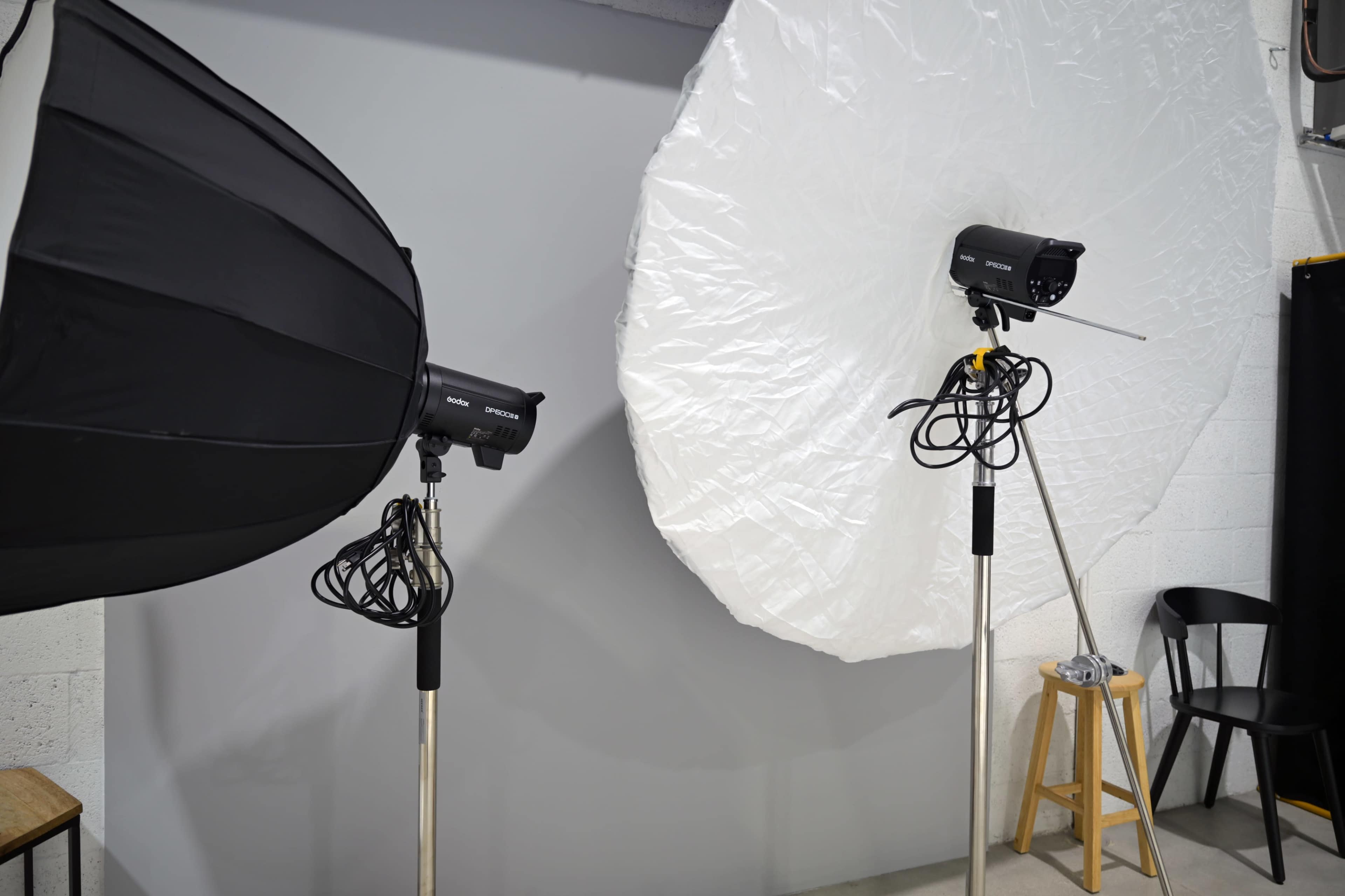 The image shows two studio lights mounted on stands, one with a black umbrella modifier and the other with a large white diffusing disk, set against a gray backdrop.