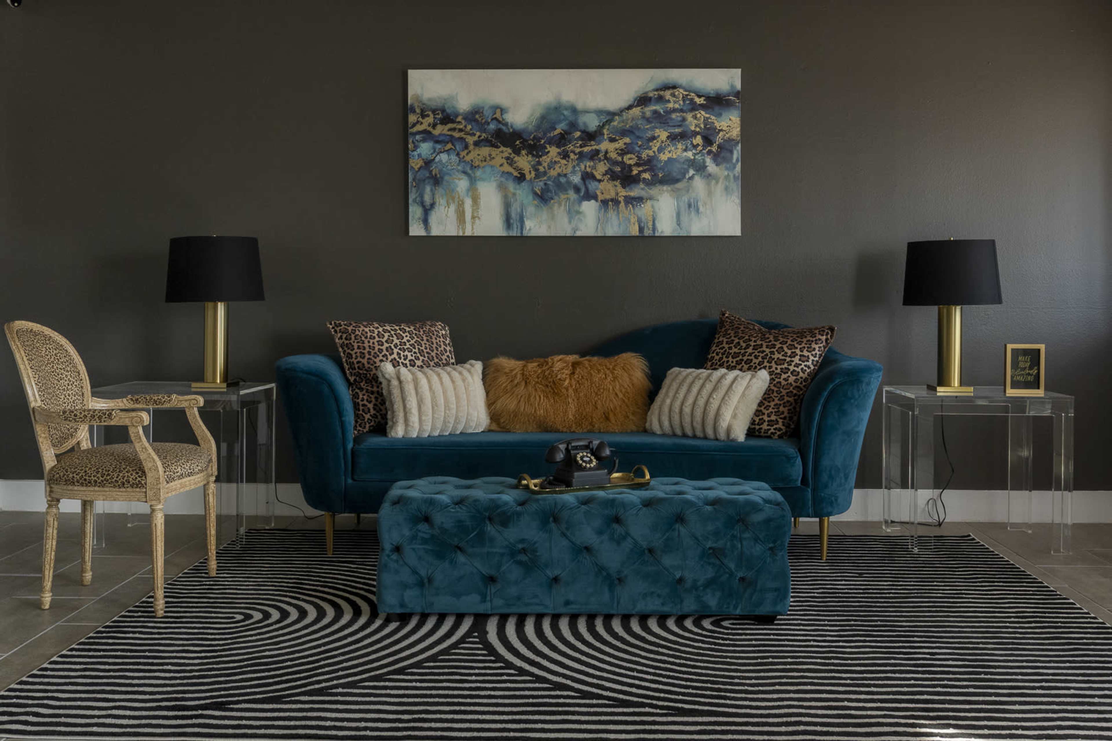 The image features a modern living room with a teal velvet sofa, two decorative throw pillows, a matching ottoman, a patterned area rug, and two black lamps on transparent side tables.