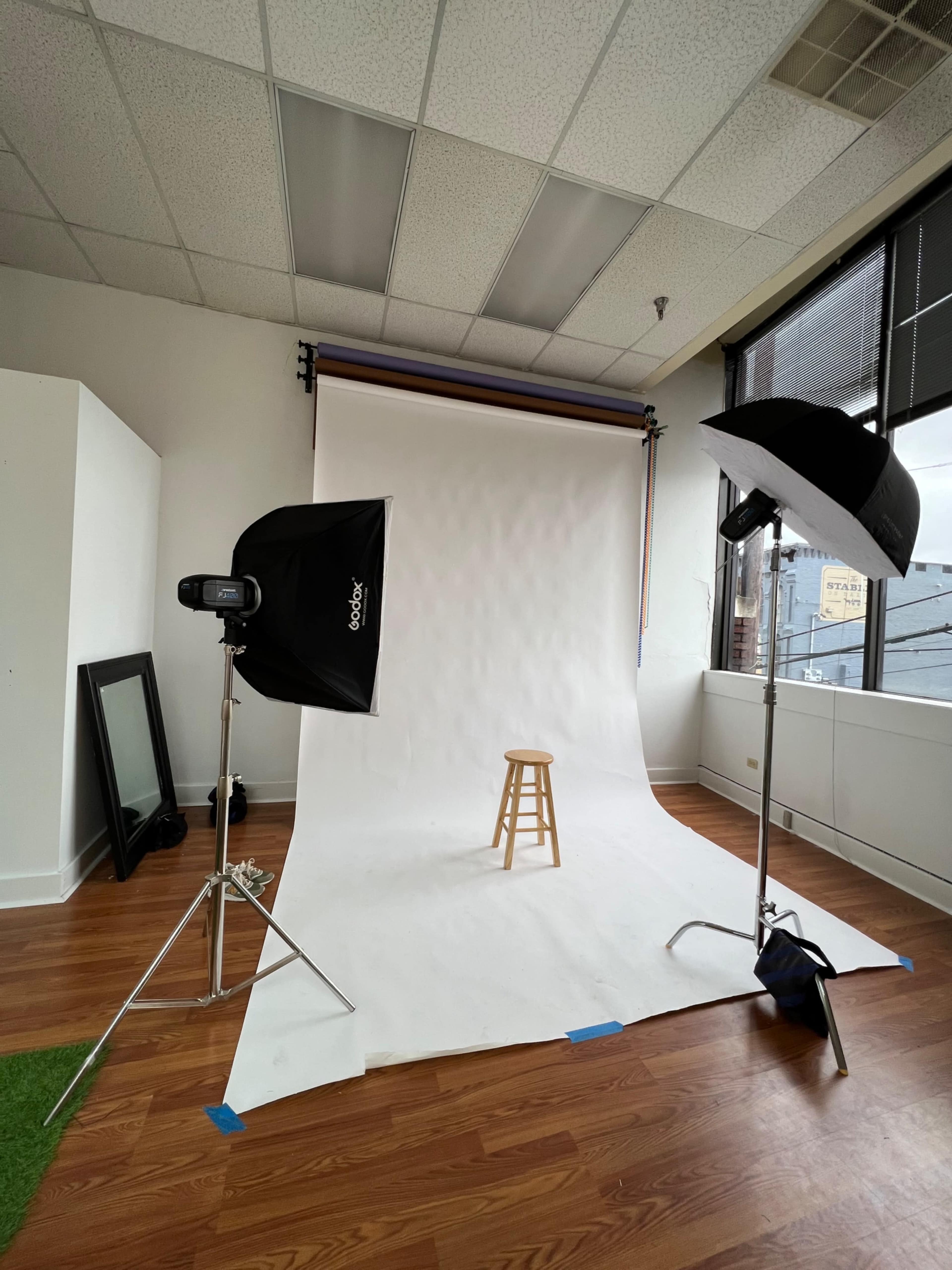A photography studio with a white backdrop, a wooden stool in the center, and two light sources positioned on either side.