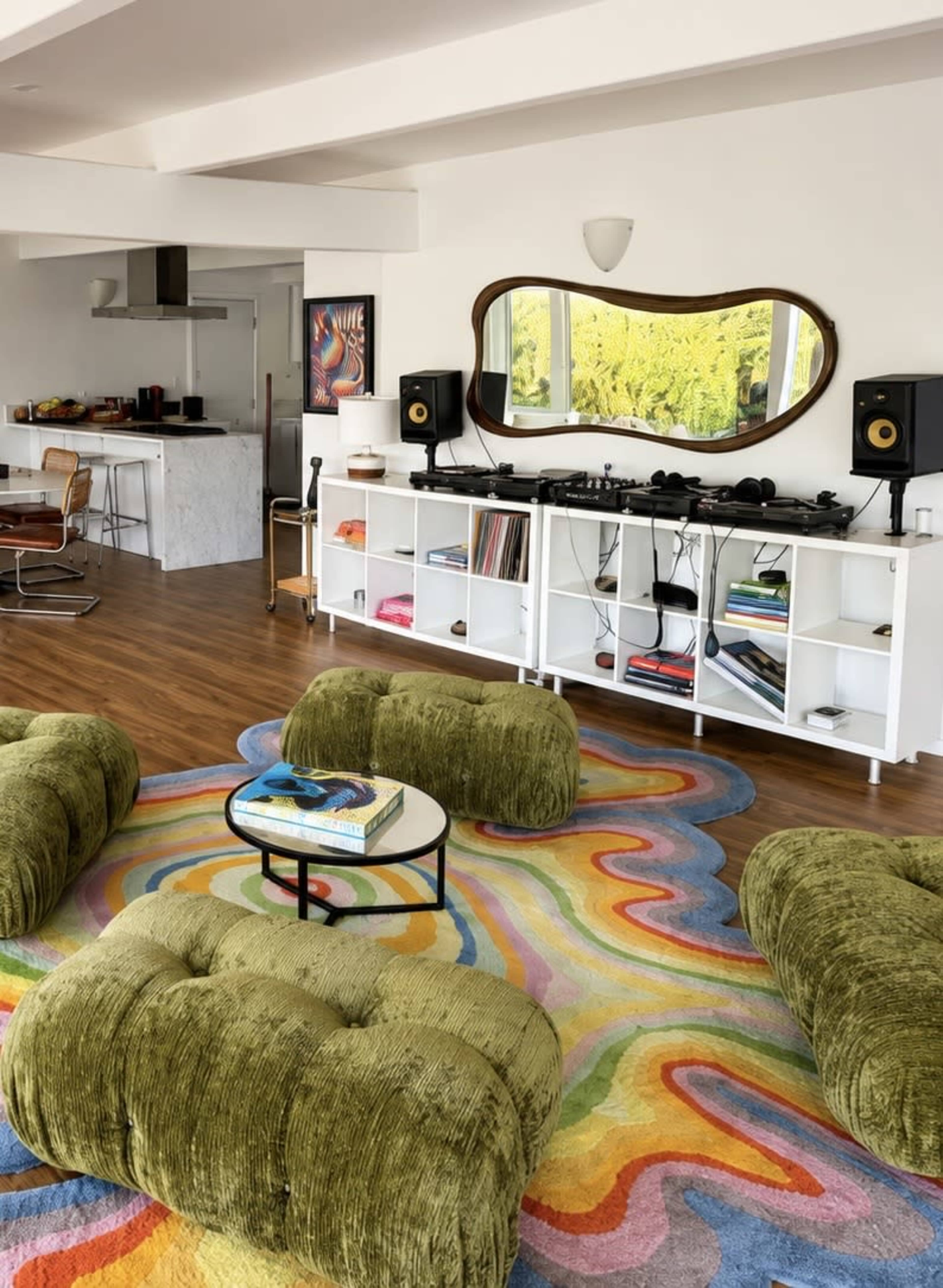 The image shows a modern living space featuring a colorful area rug, round green upholstered seating, and a white shelving unit with audio equipment beside a kitchen area.