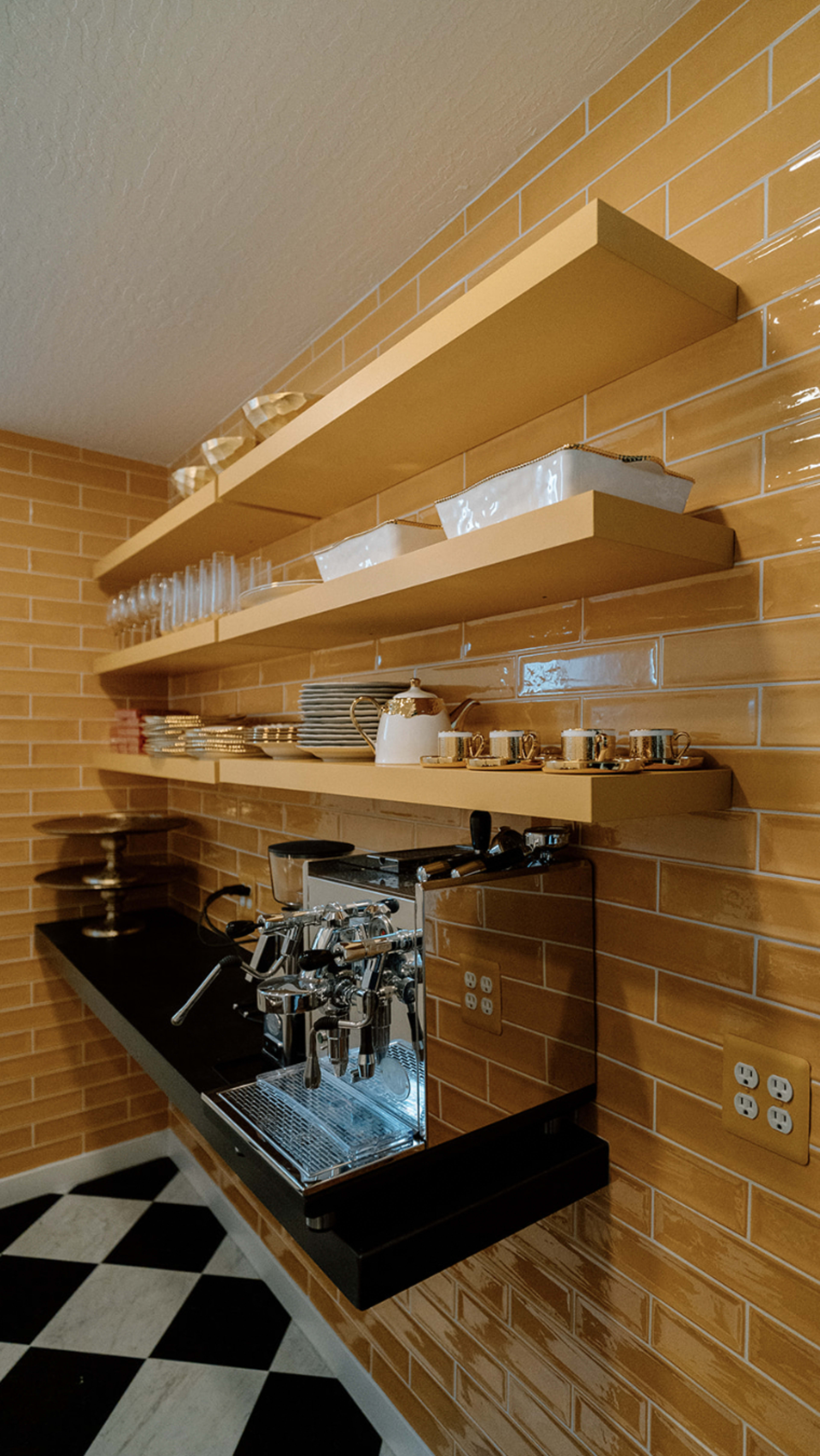 A kitchen features a coffee machine on a black countertop, with yellow tile walls and open shelves displaying glassware and dishware.