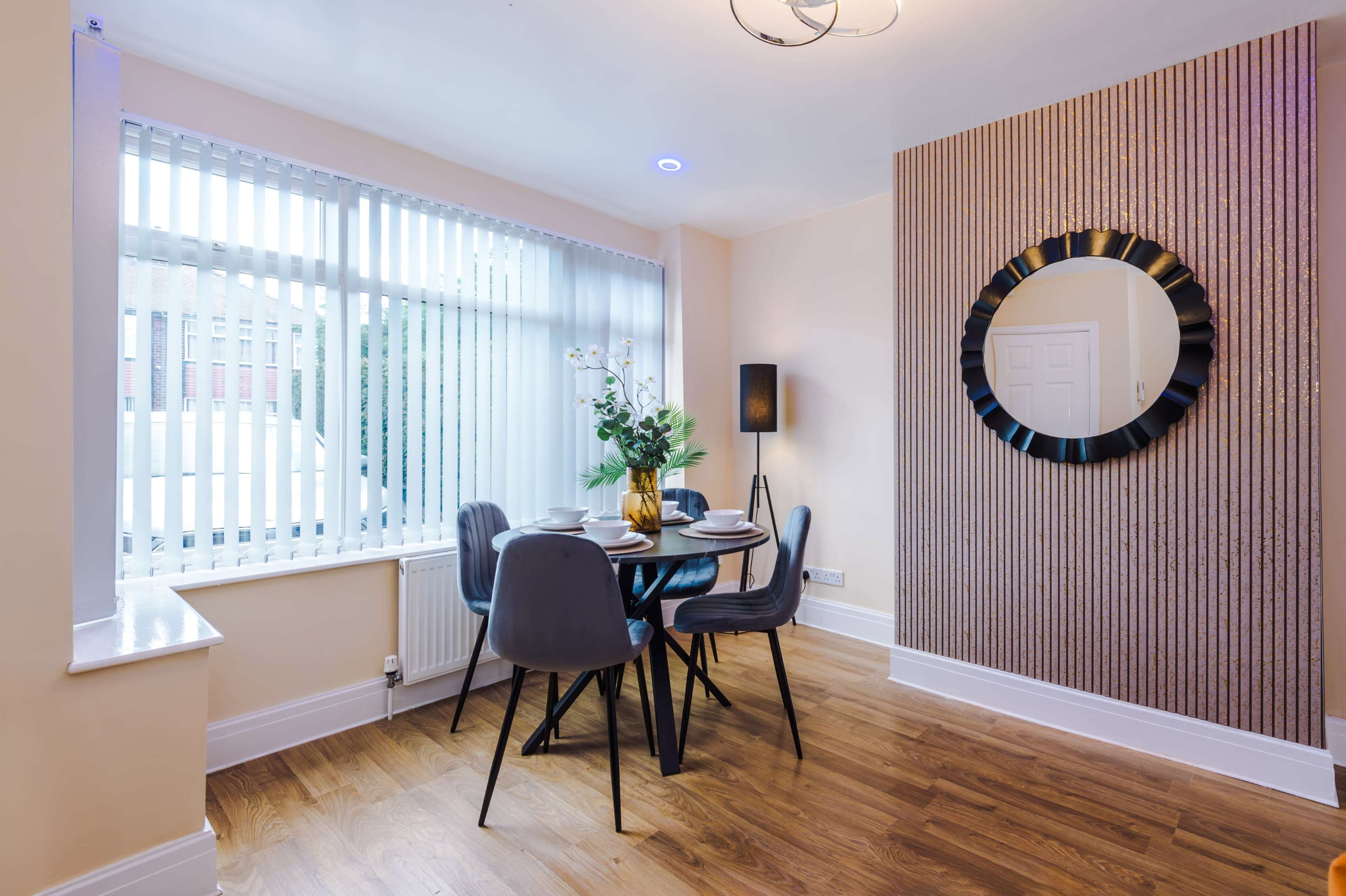 The room features a dining area with a round table, four chairs, and large windows adorned with vertical blinds.