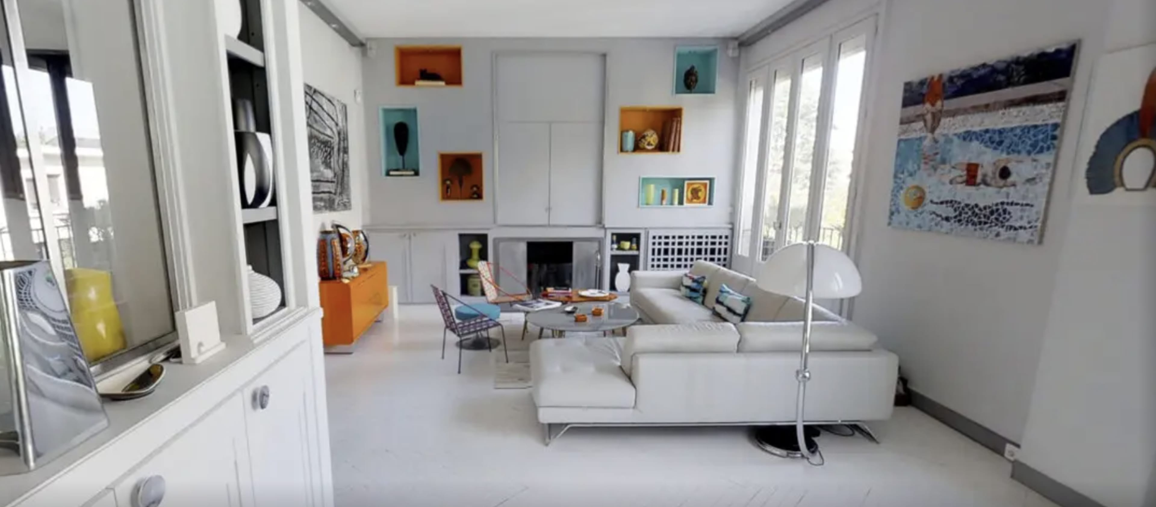 A modern living room featuring a white leather sofa, a glass coffee table, and colorful wall art, with large windows allowing ample natural light.