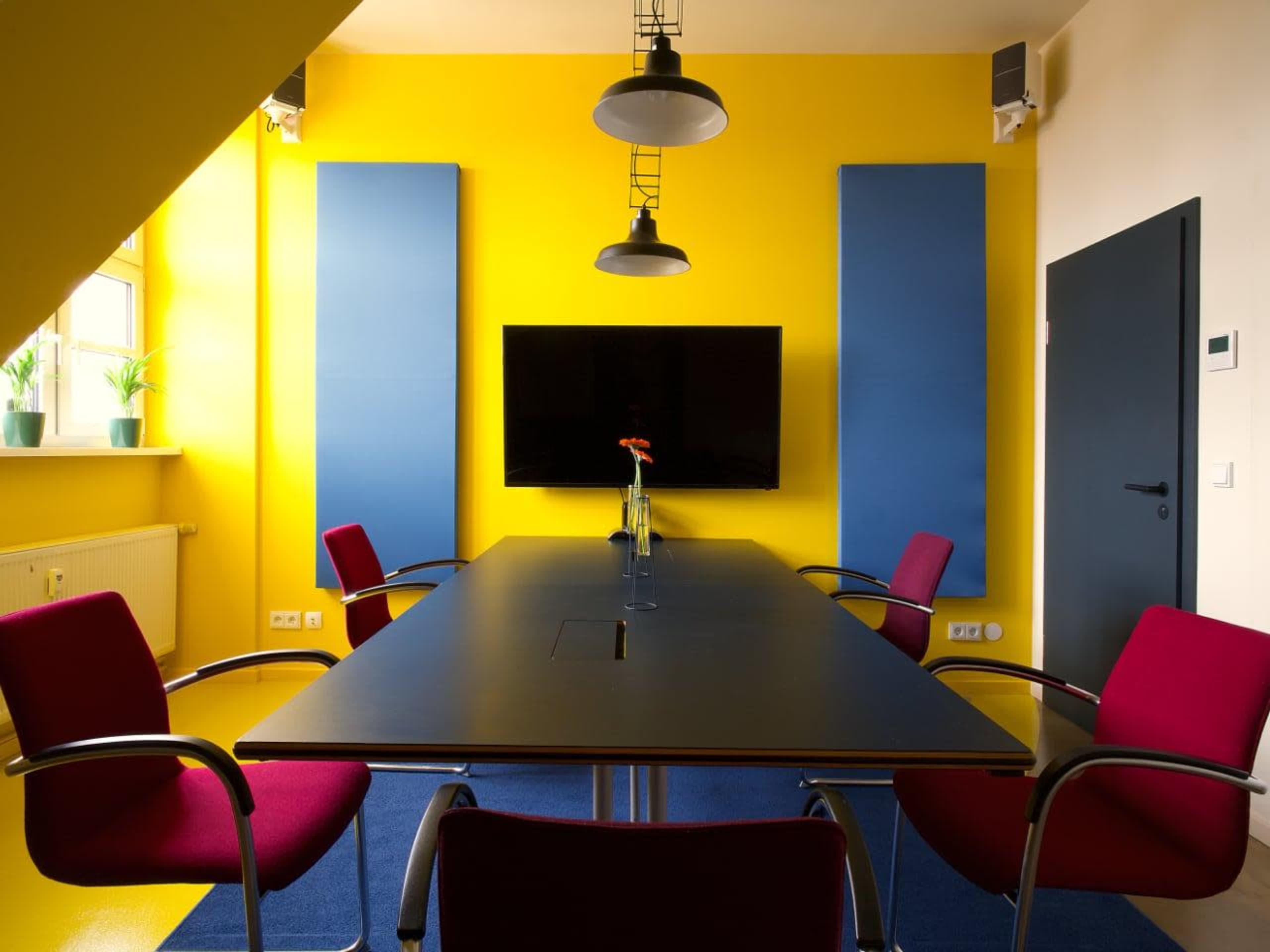 A modern conference room features a black table surrounded by red chairs, a large screen on the wall, and bright yellow walls with sound-absorbing panels.