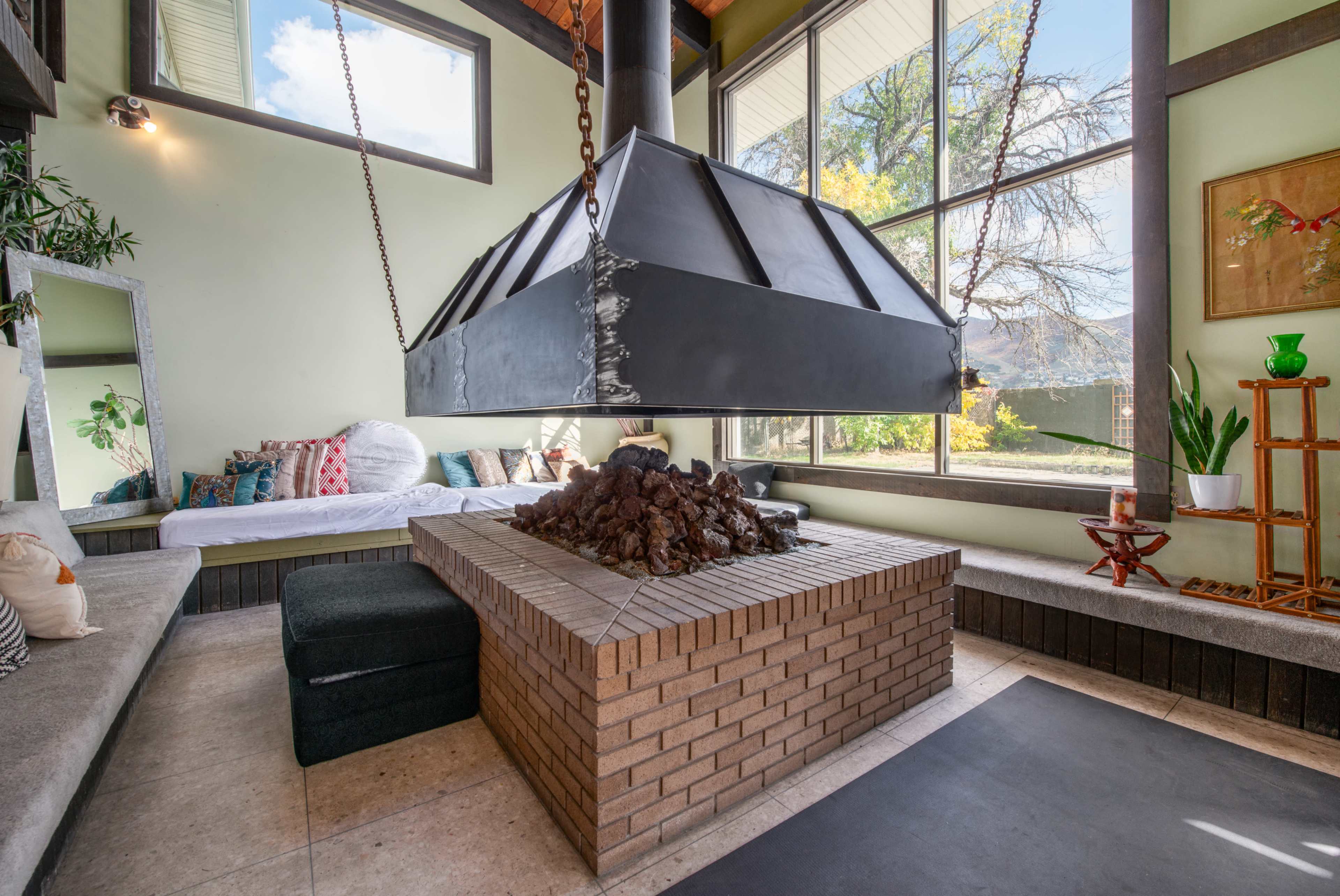 A modern lounge area featuring a central fireplace with a hanging metal hood, surrounded by seating and large windows that let in natural light.