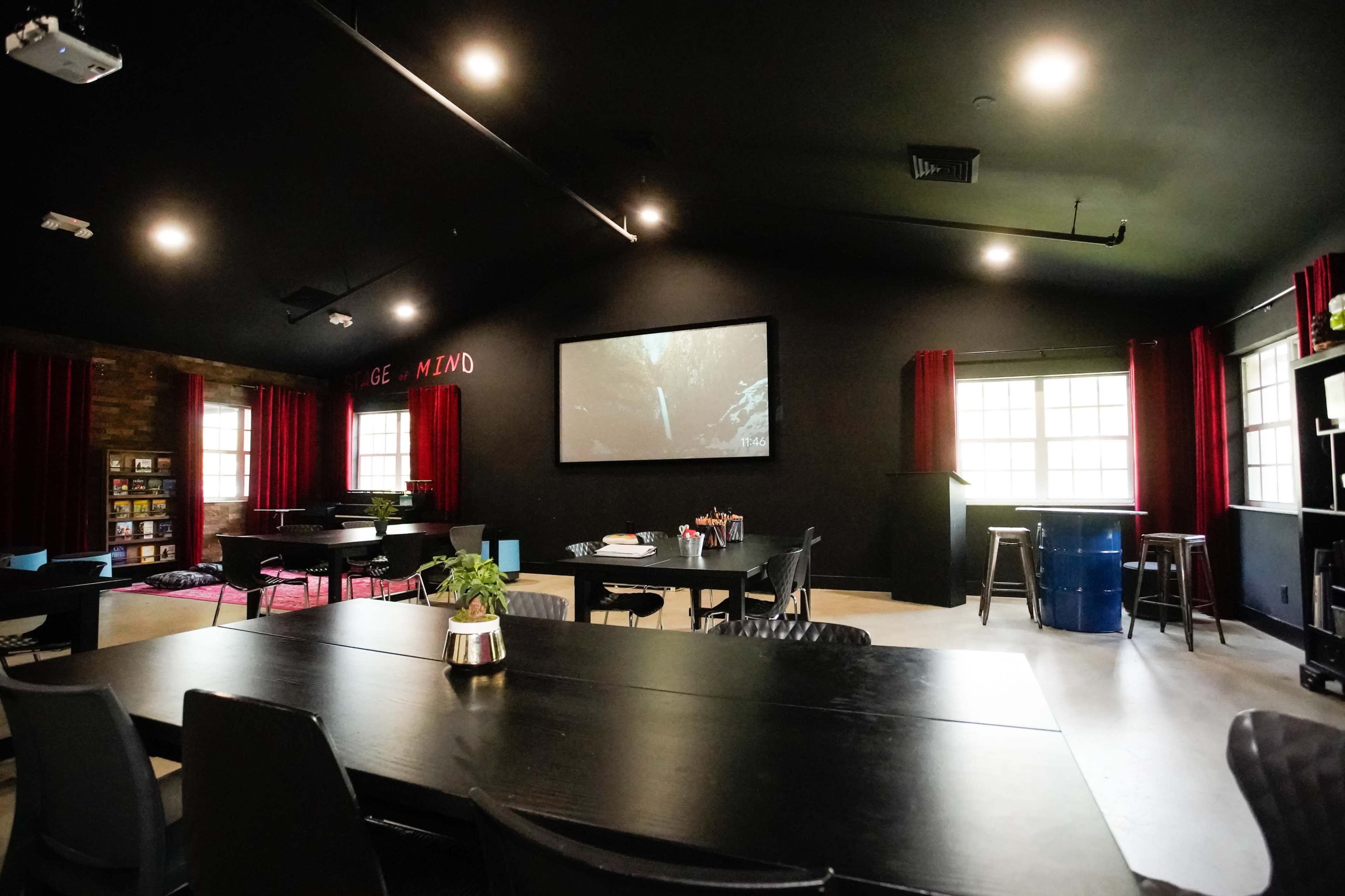 The image shows a modern meeting room with a large screen at the front, red curtains, and several tables and chairs arranged for collaboration.