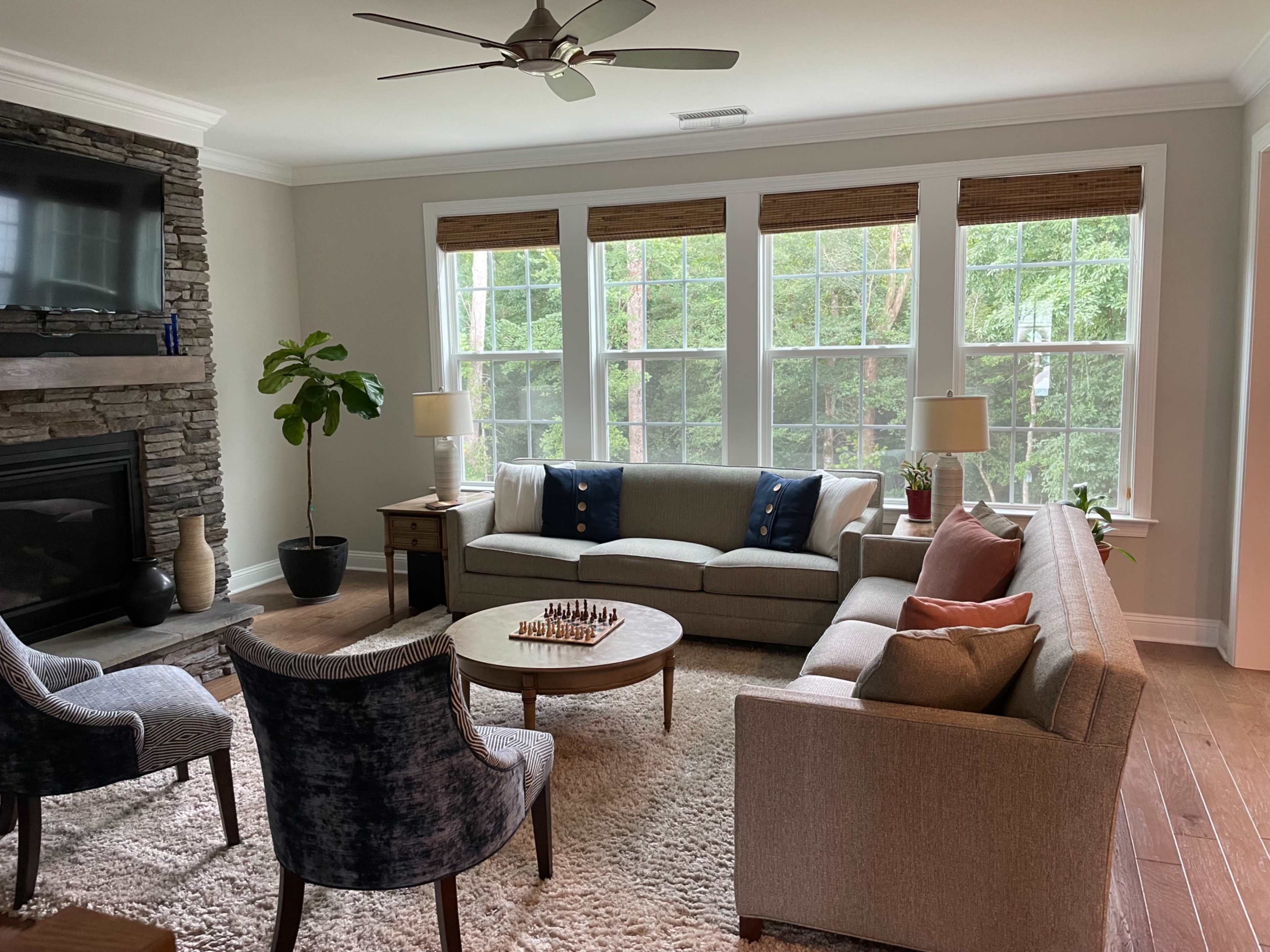 A light-filled living room features a gray sofa, two upholstered armchairs, a round coffee table with a chess set, and large windows overlooking greenery.