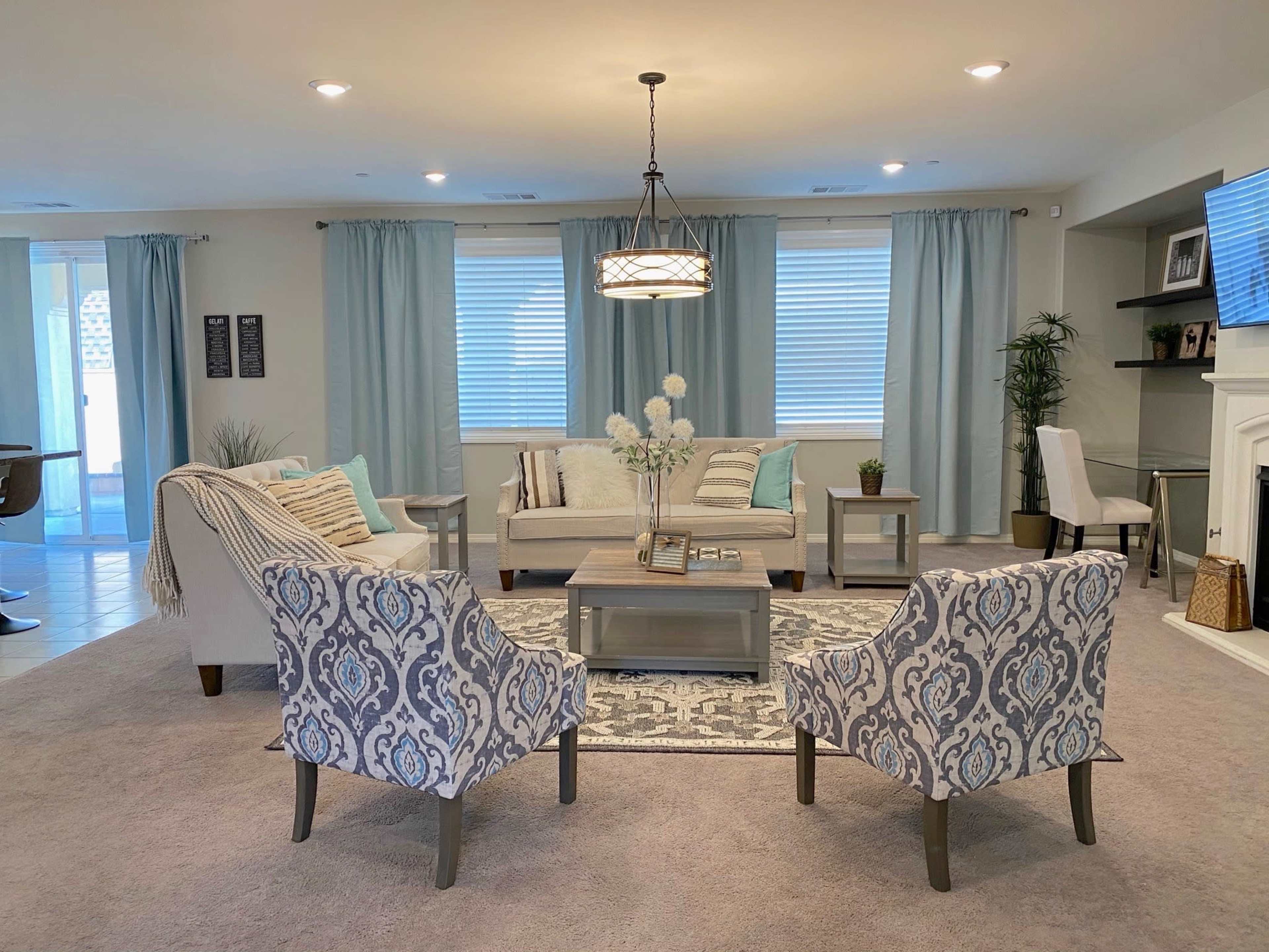 A cozy living room features a central rug with a coffee table surrounded by patterned chairs and sofas, with light curtains framing the windows.