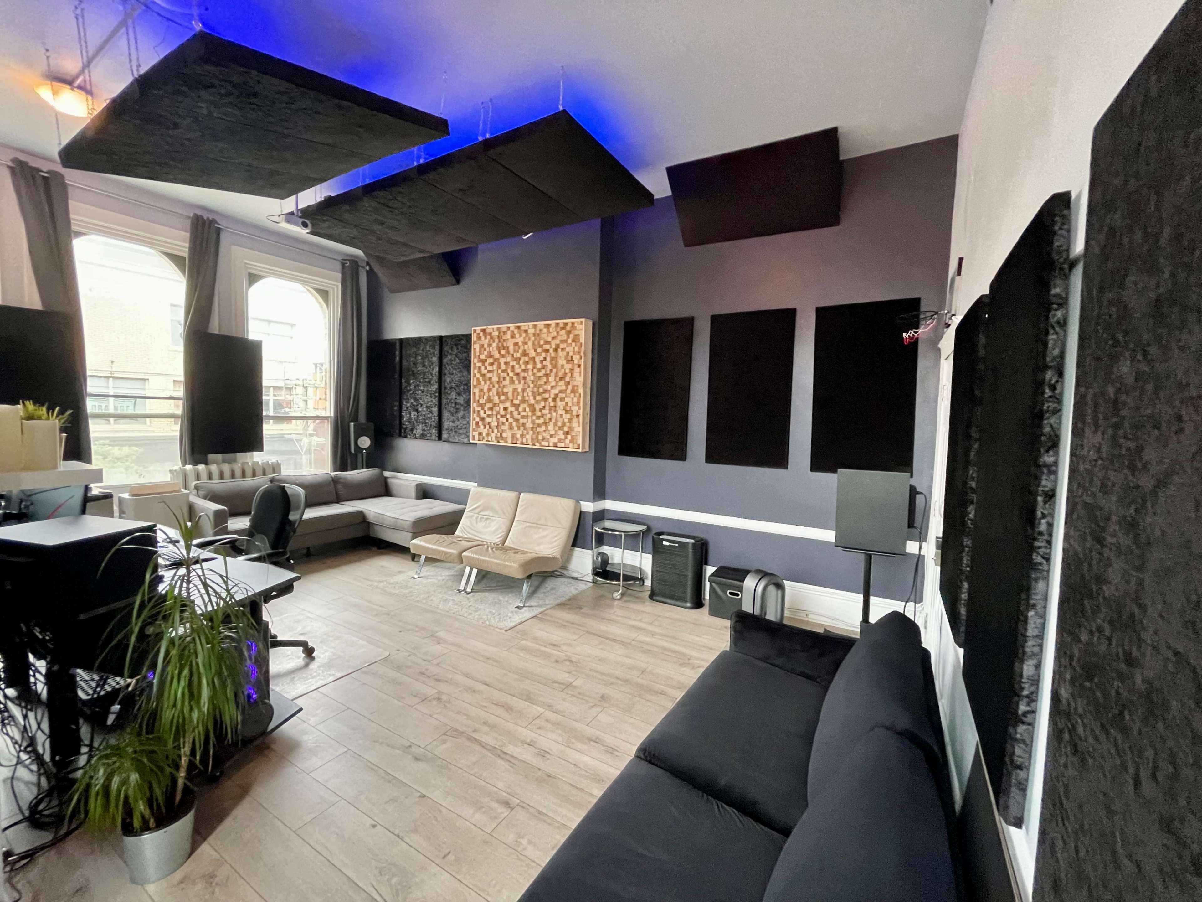 A modern studio space with soundproofing panels on the walls, a mix of seating options including a sofa and a futon, and a desk with a plant in the foreground.