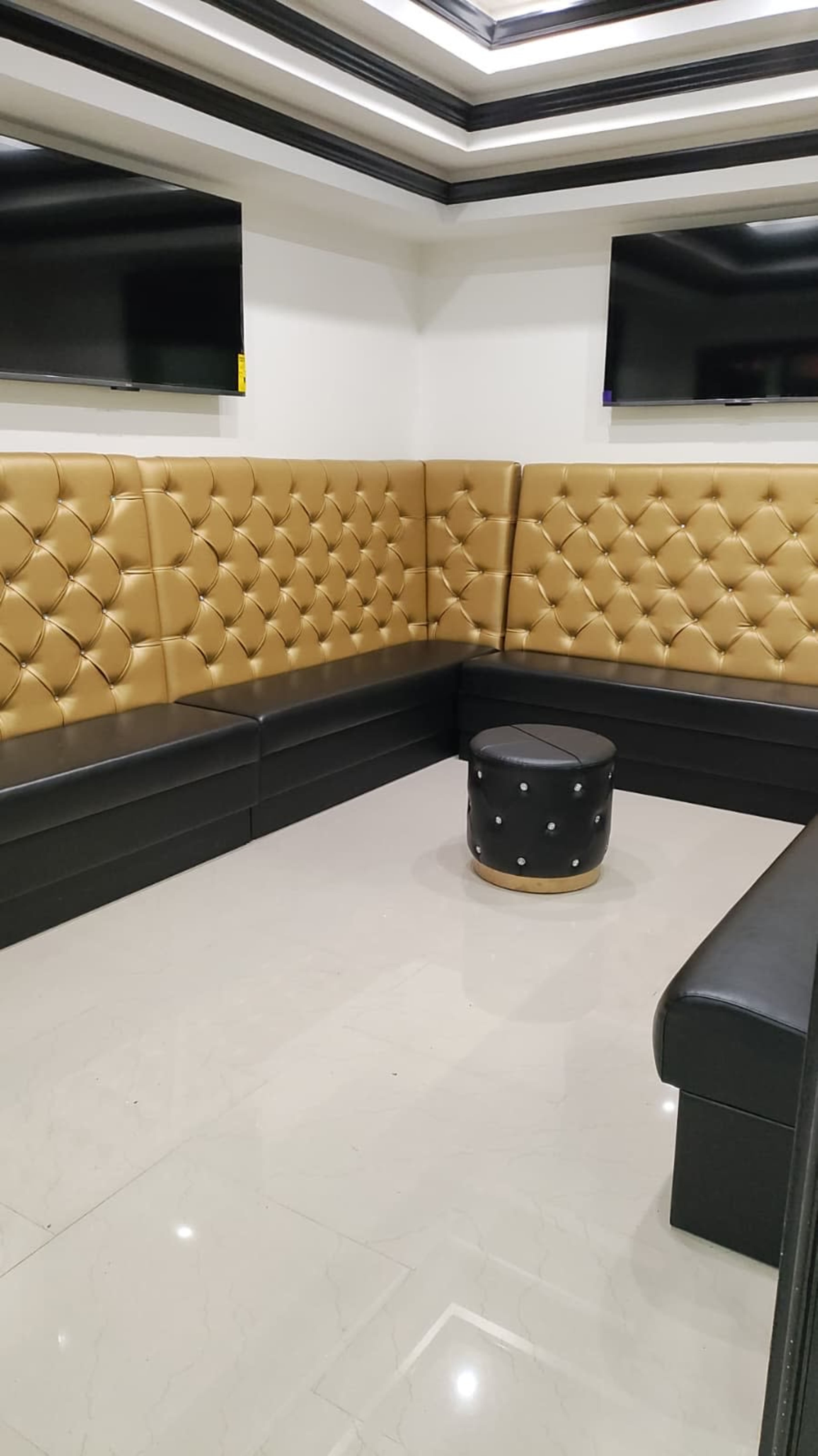The image shows a small lounge area with black and gold tufted seating, two flat-screen TVs mounted on the wall, and a round black stool in the center.