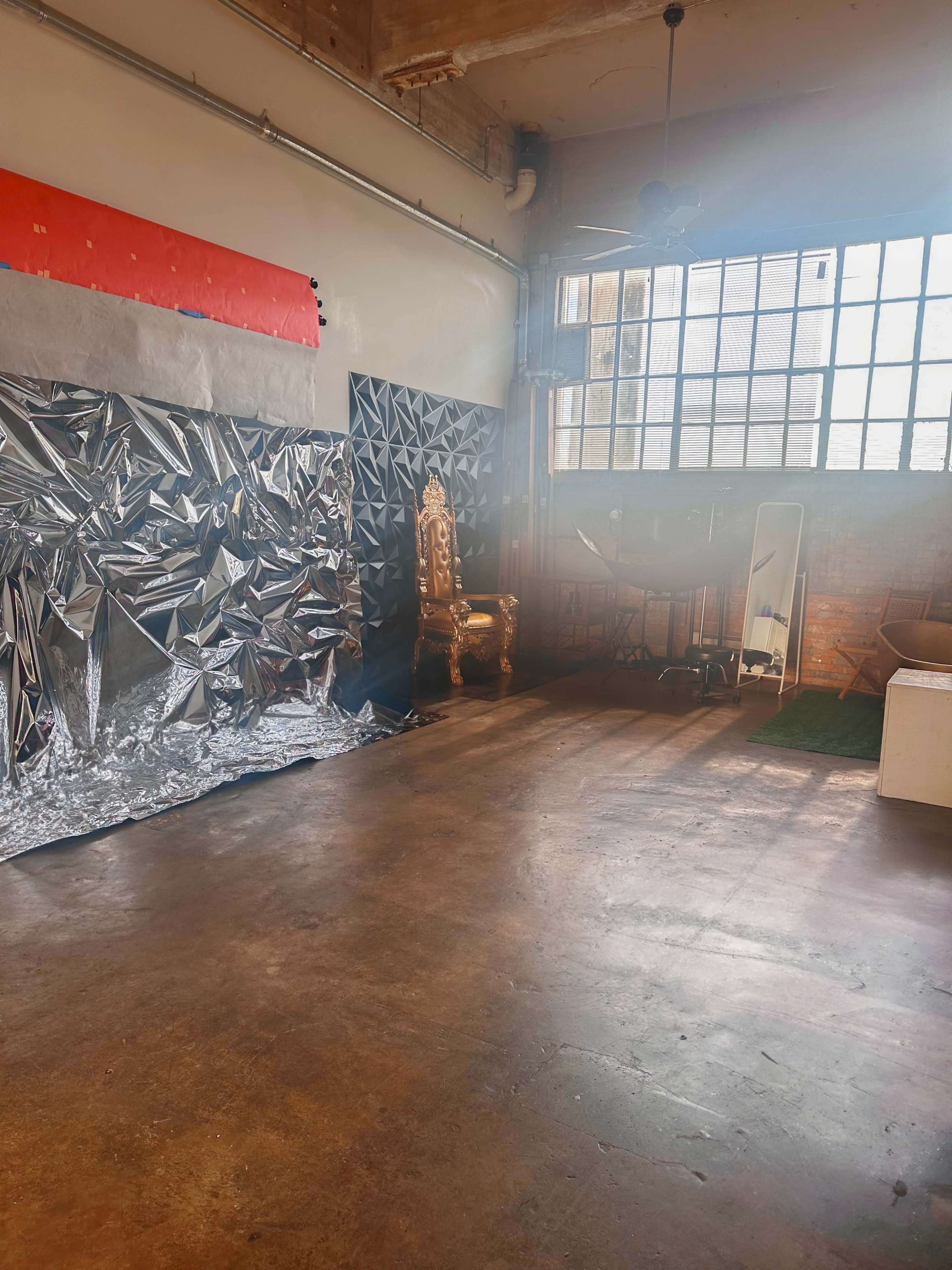 The image shows an industrial-style room with a large window, a golden chair, and a wall covered in reflective silver material.