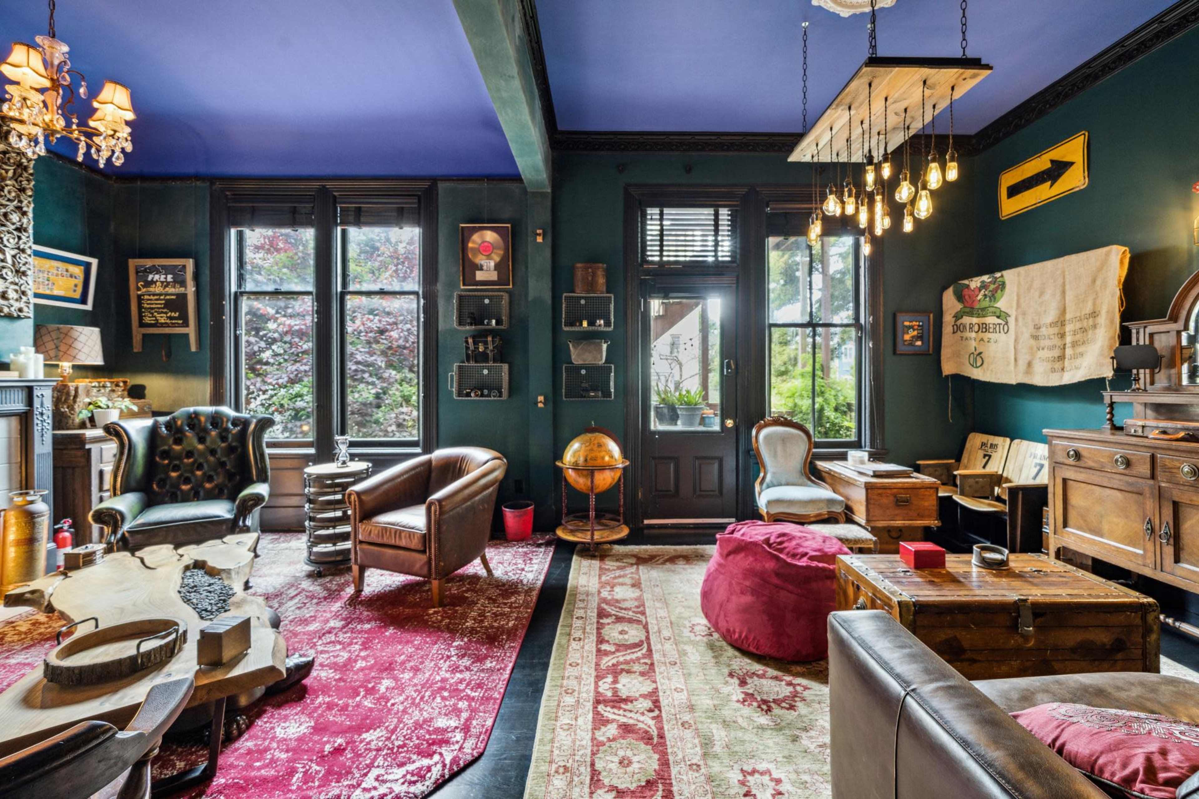 A cozy room with dark green walls, a blue ceiling, various seating options, wooden furniture, and large windows letting in natural light.
