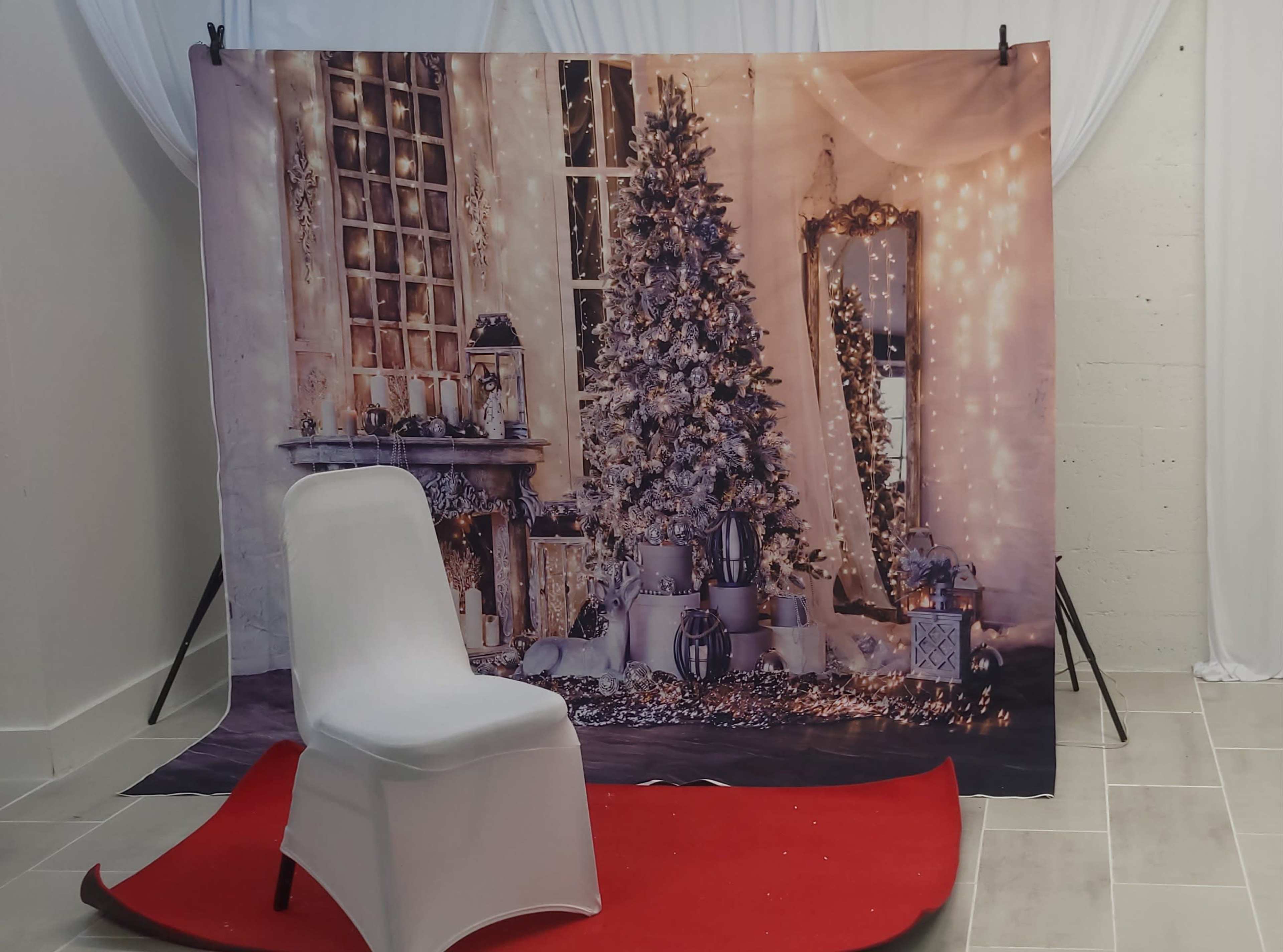 A white chair is positioned in front of a backdrop featuring a decorated Christmas tree and a mirror in a softly lit, festive setting.