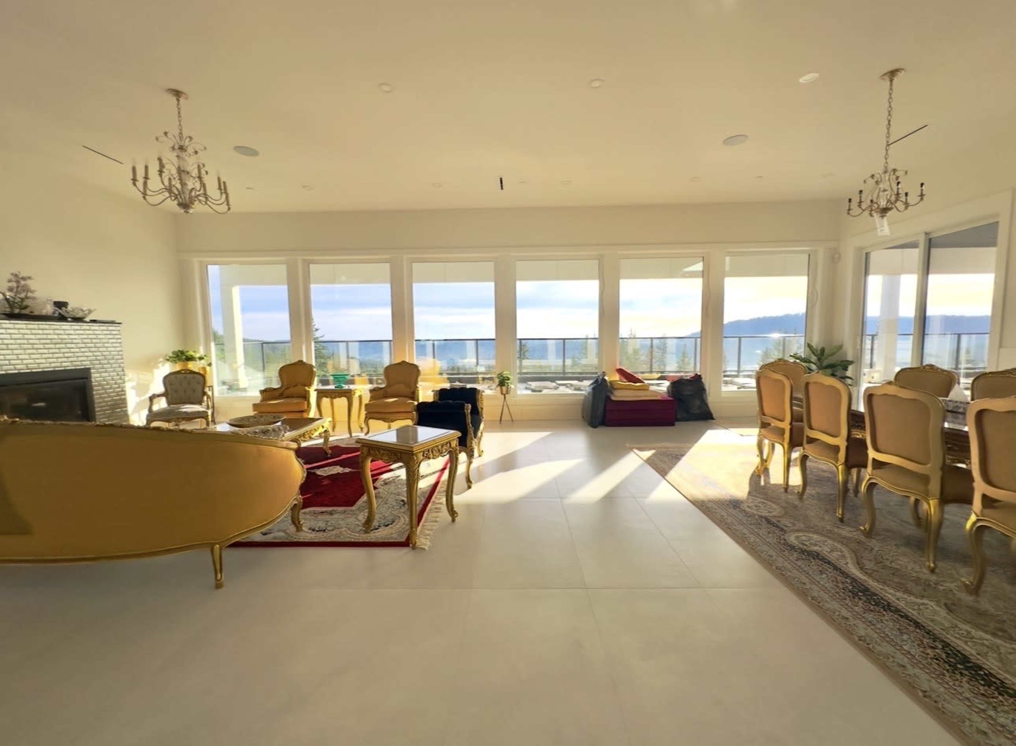 The image shows a spacious living room with large windows overlooking a body of water, featuring elegant furniture including a gold-accented sofa and dining chairs.