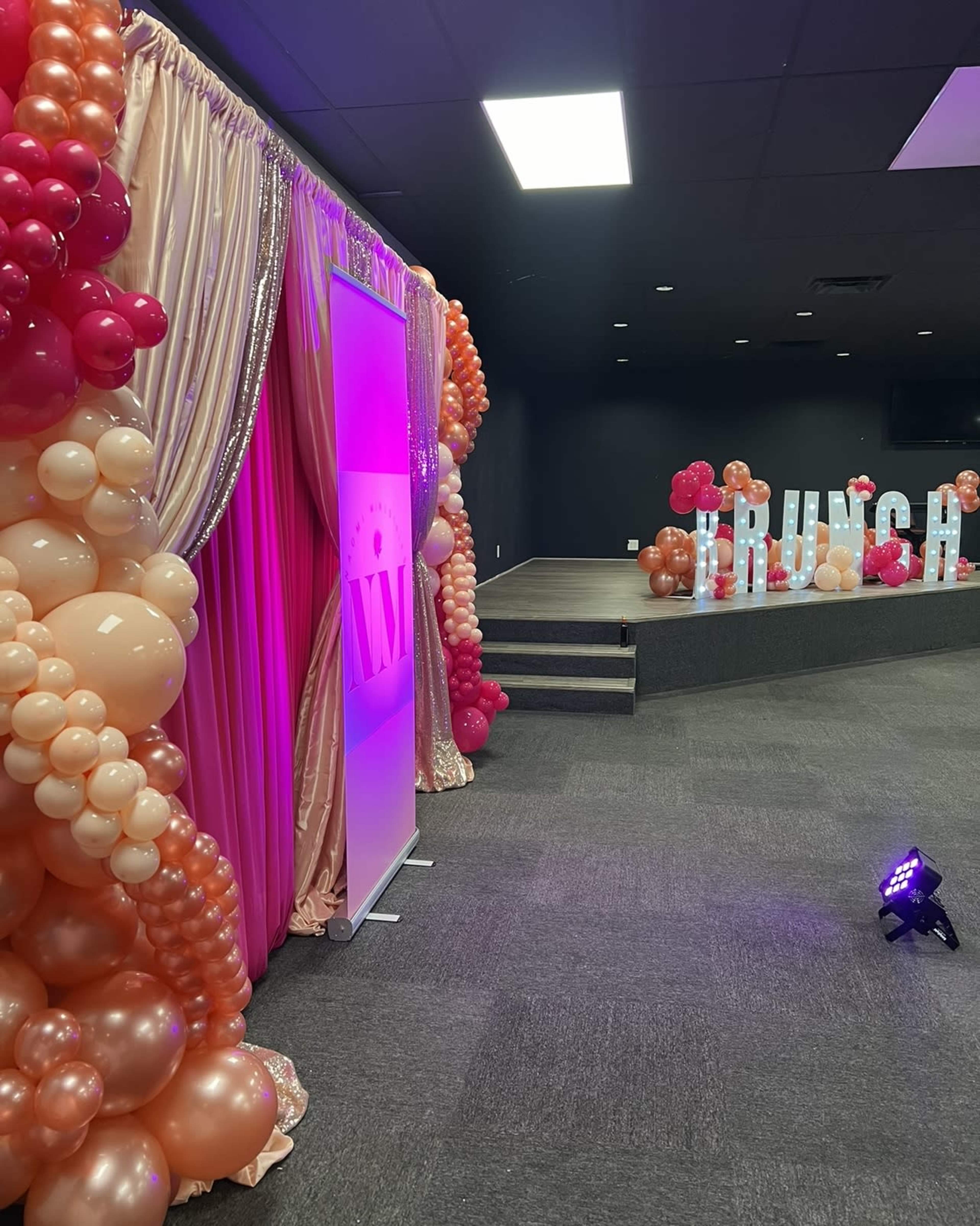 The image shows a decorated event space featuring a backdrop with draped fabric and balloons in shades of pink and peach, along with large letters spelling "BRUNCH" on a raised platform.