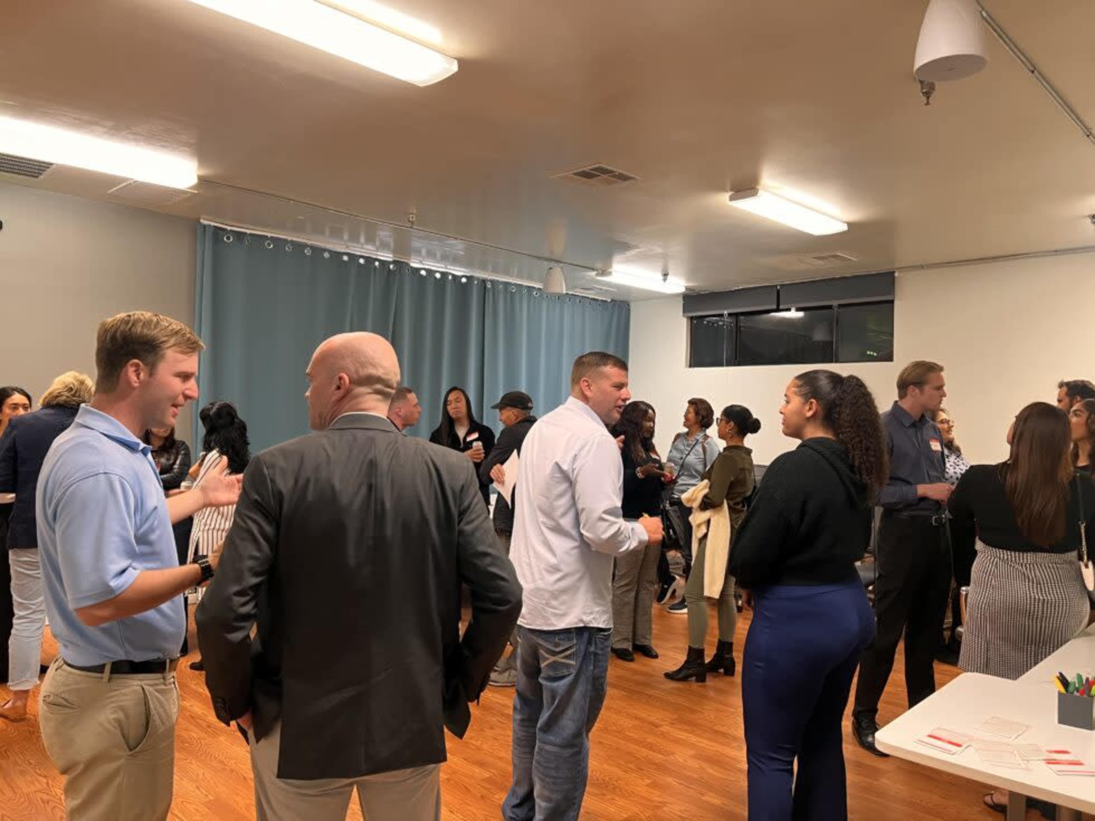 A large group of people engage in conversation at a social event in a well-lit room with wooden floors.