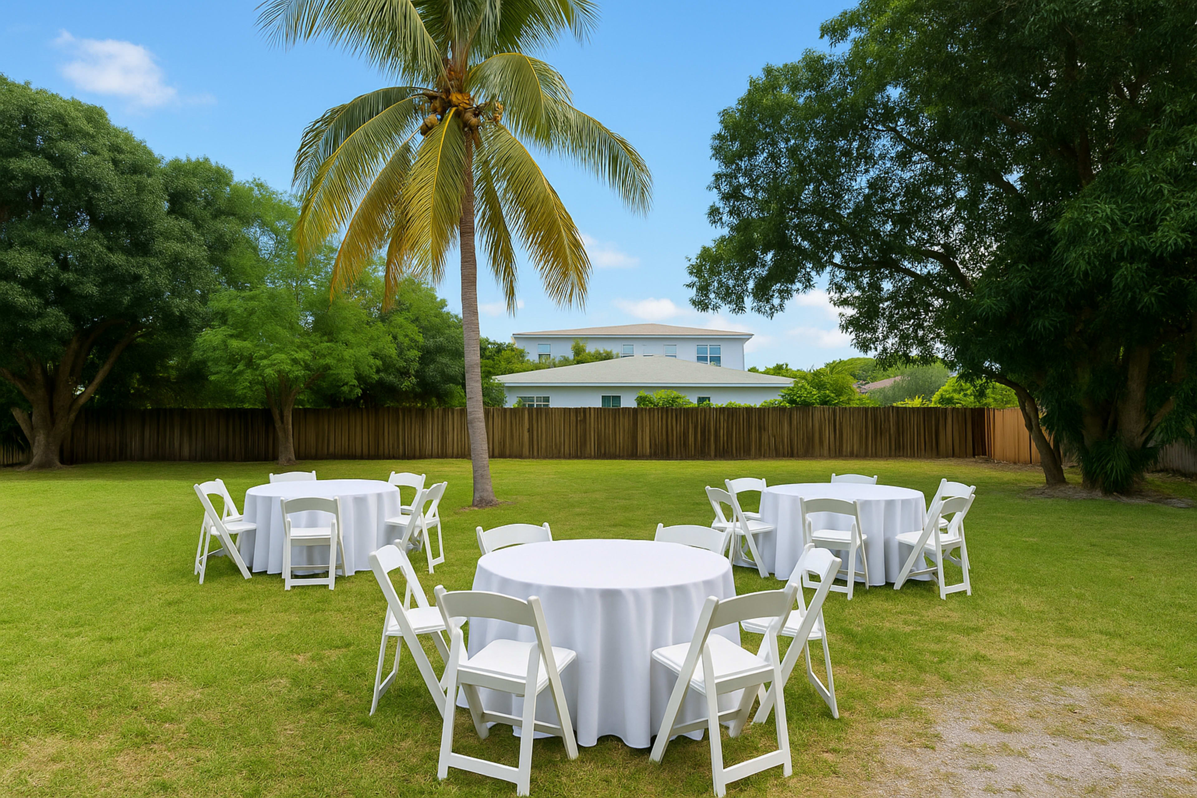 A grassy area features circular tables with white tablecloths surrounded by white chairs, with palm trees and a house in the background.