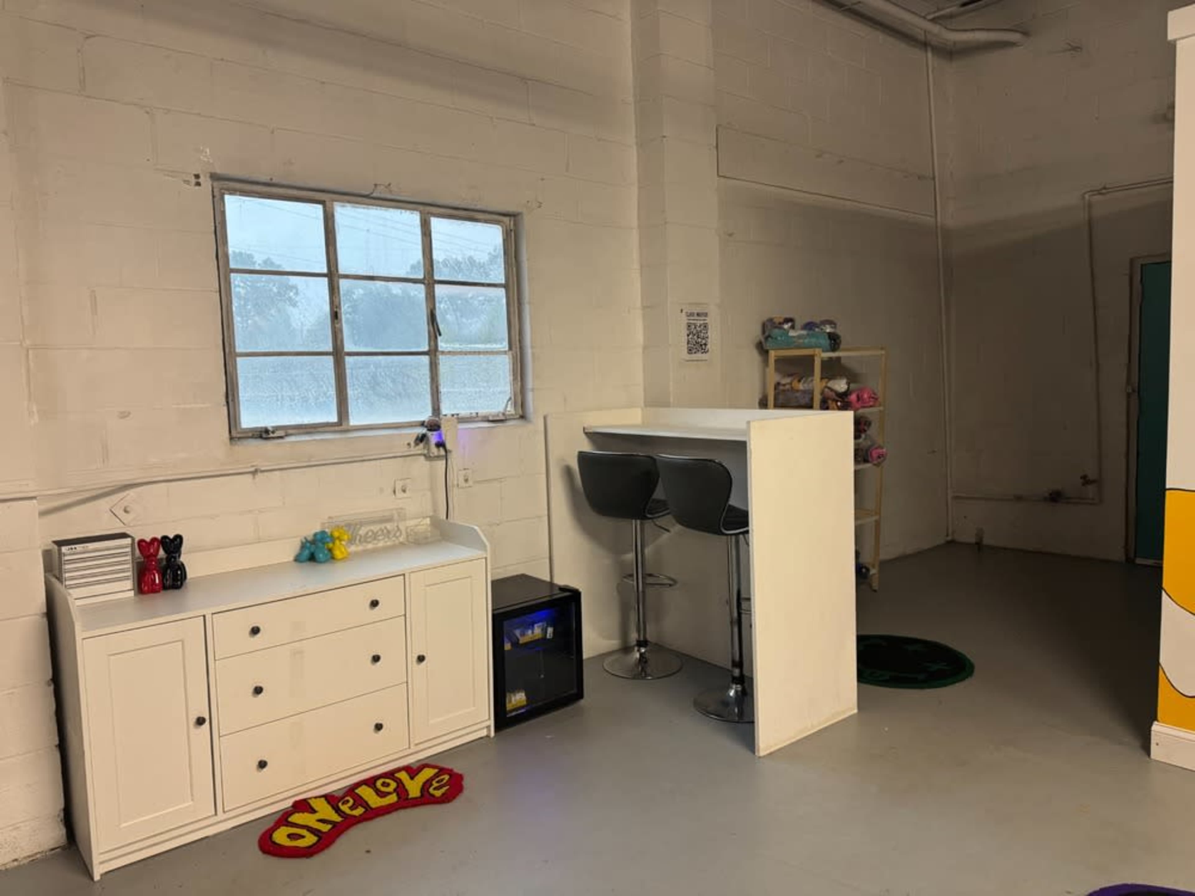 The image shows a bright, minimalist indoor space featuring a white dresser, a small bar table with stools, and a shelf with colorful plush toys next to a window.