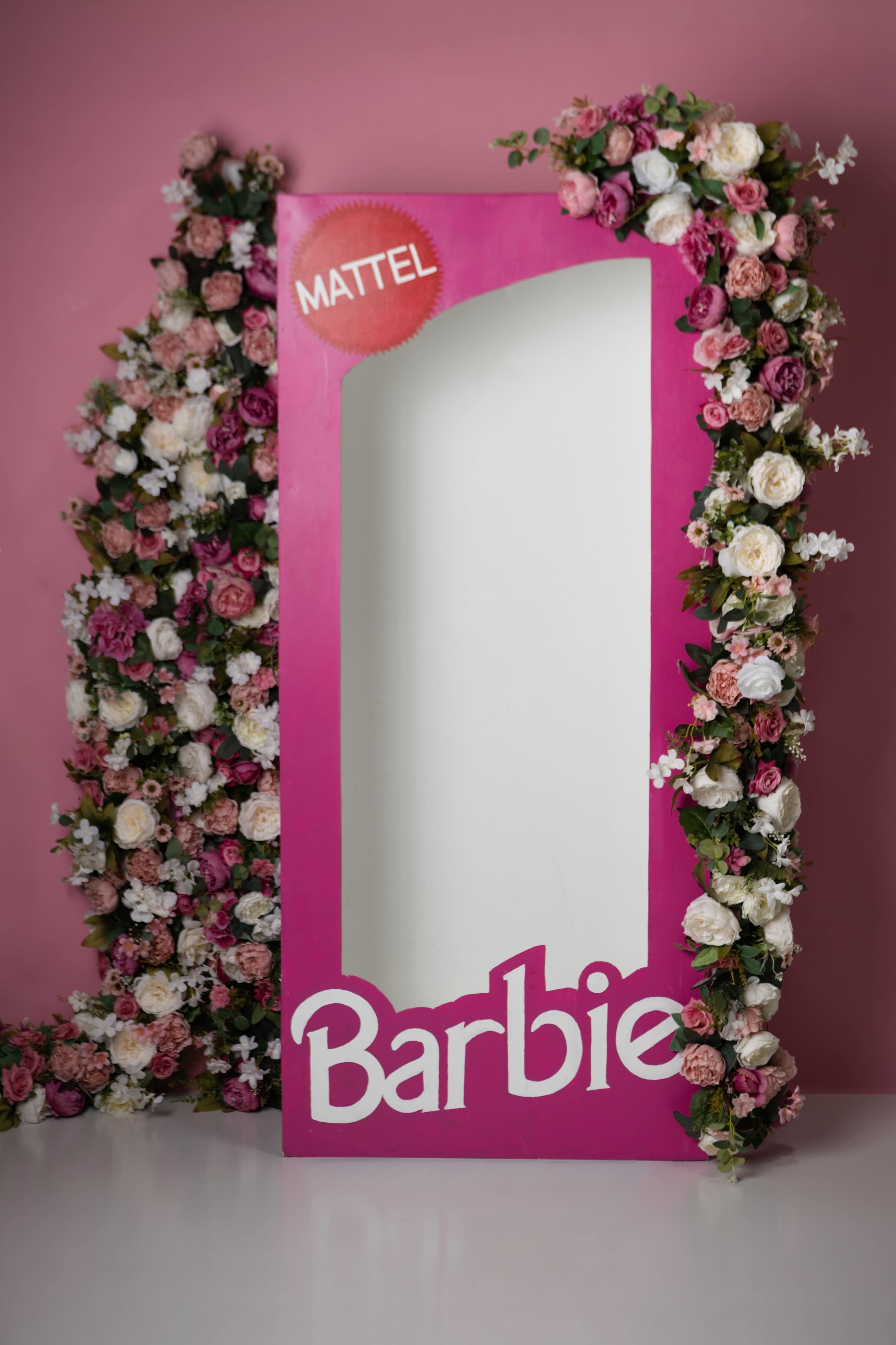The image features a large, pink frame adorned with artificial flowers, prominently displaying the word "Barbie" and a Mattel logo at the top.