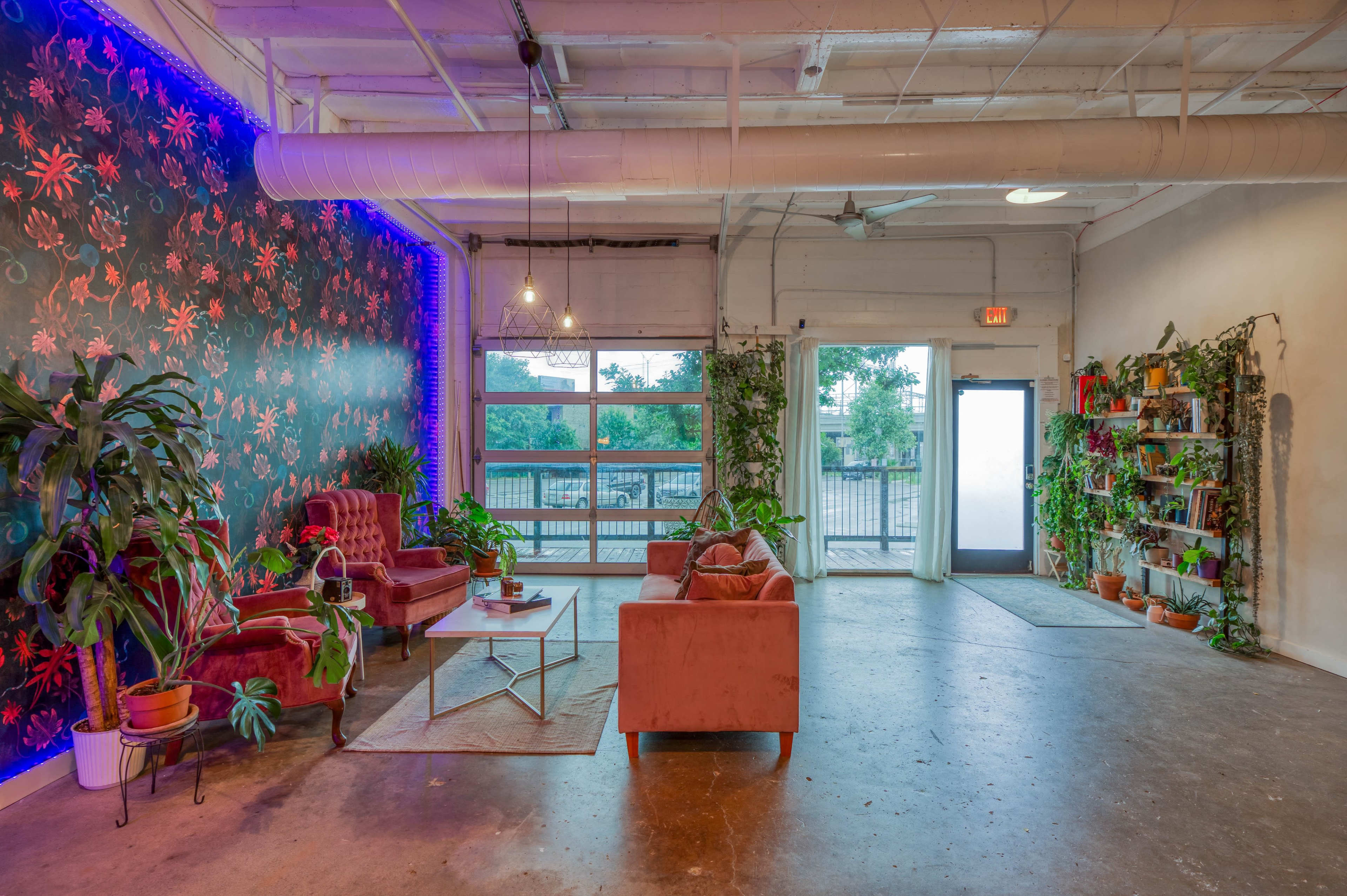 The room features two red sofas arranged around a coffee table, with a wall decorated with floral wallpaper and various plants positioned throughout the space.