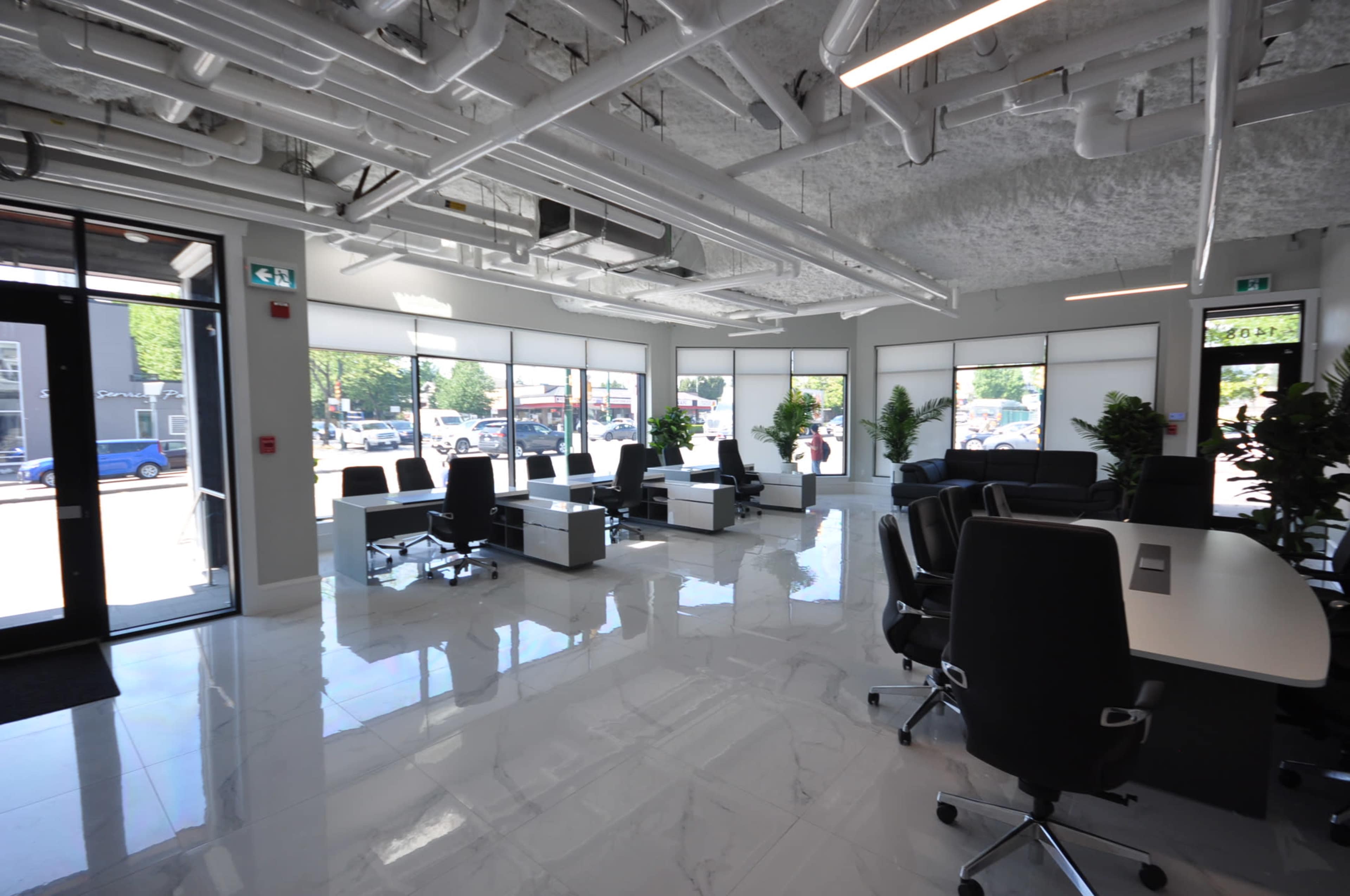 A modern office space with white walls, large windows, and multiple workstations arranged with black office chairs.