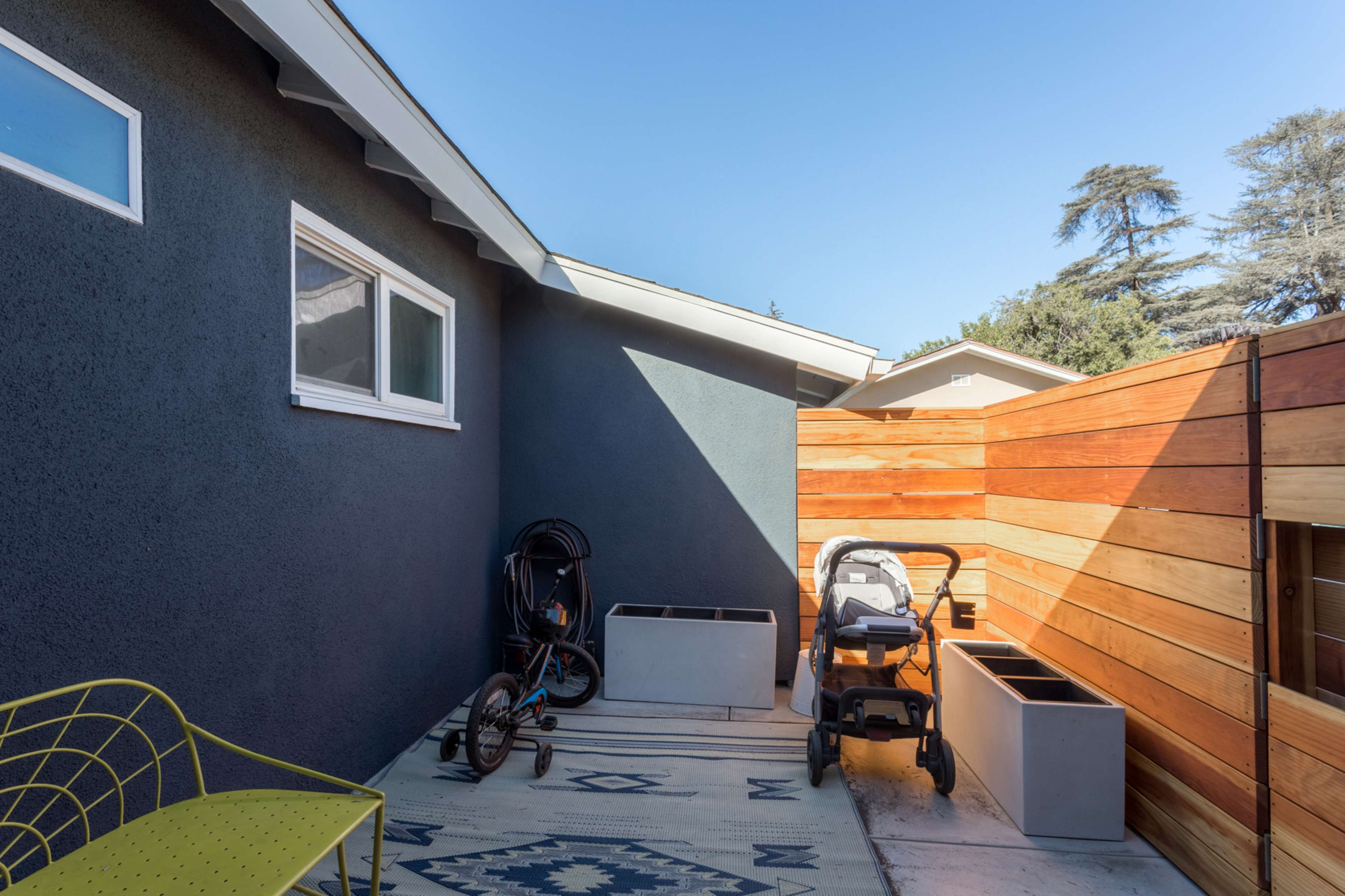 A small outdoor patio features a bicycle, a stroller, and two planter boxes against a wooden fence.