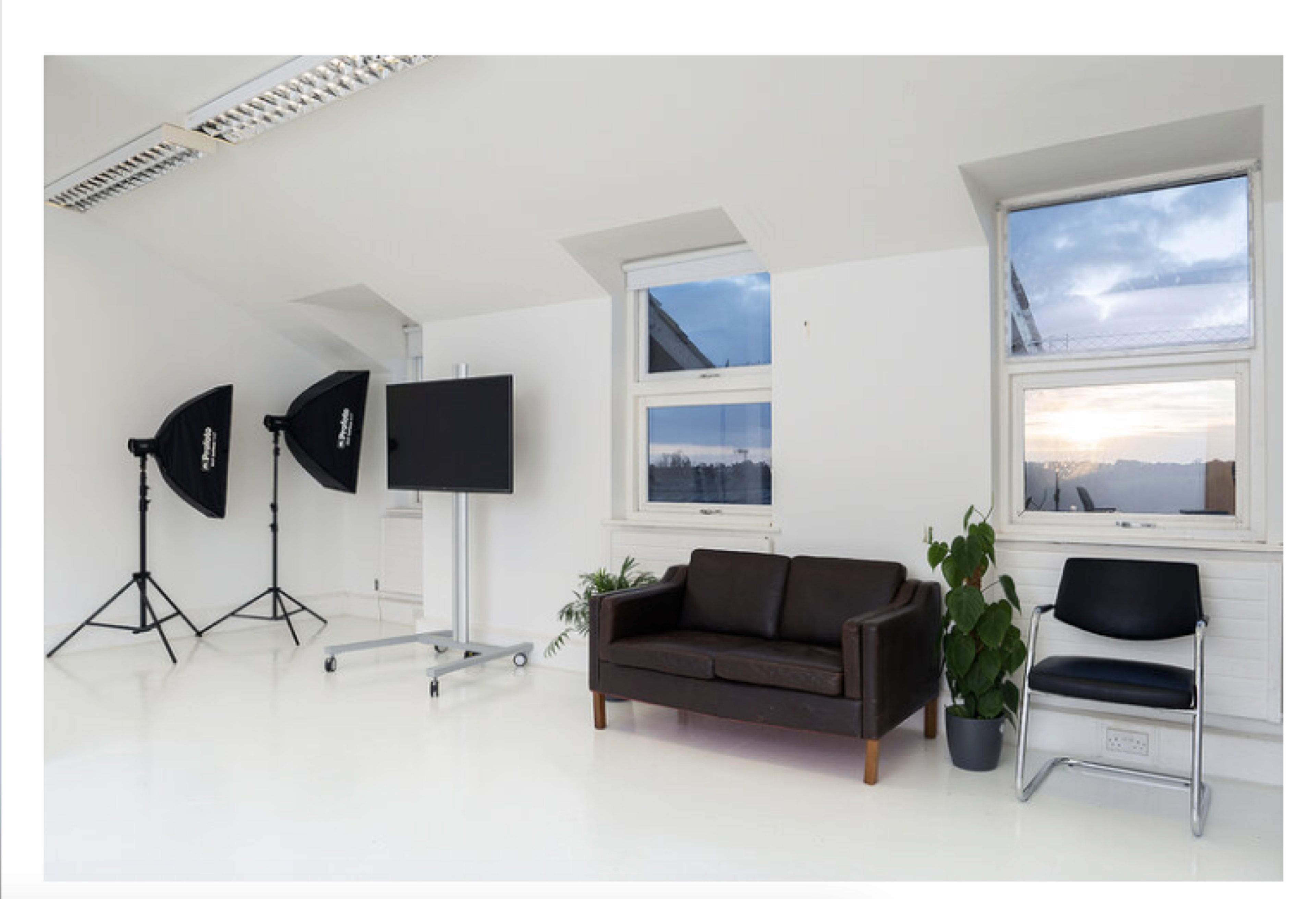 The image shows a bright studio space with a brown leather sofa, a black chair, a large screen on a mobile stand, and two lighting setup umbrellas.