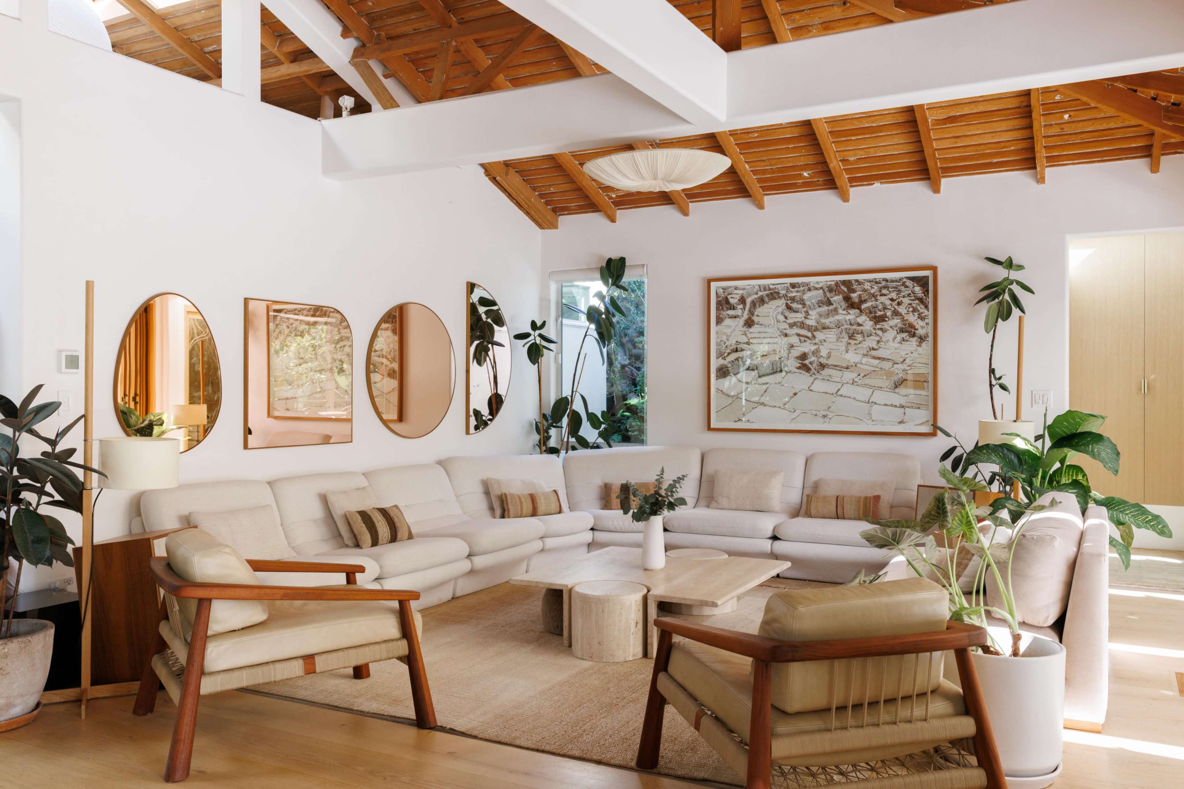 A bright living room features a large sectional sofa, a wooden coffee table, and multiple mirrors on the wall, surrounded by indoor plants and natural wood accents.