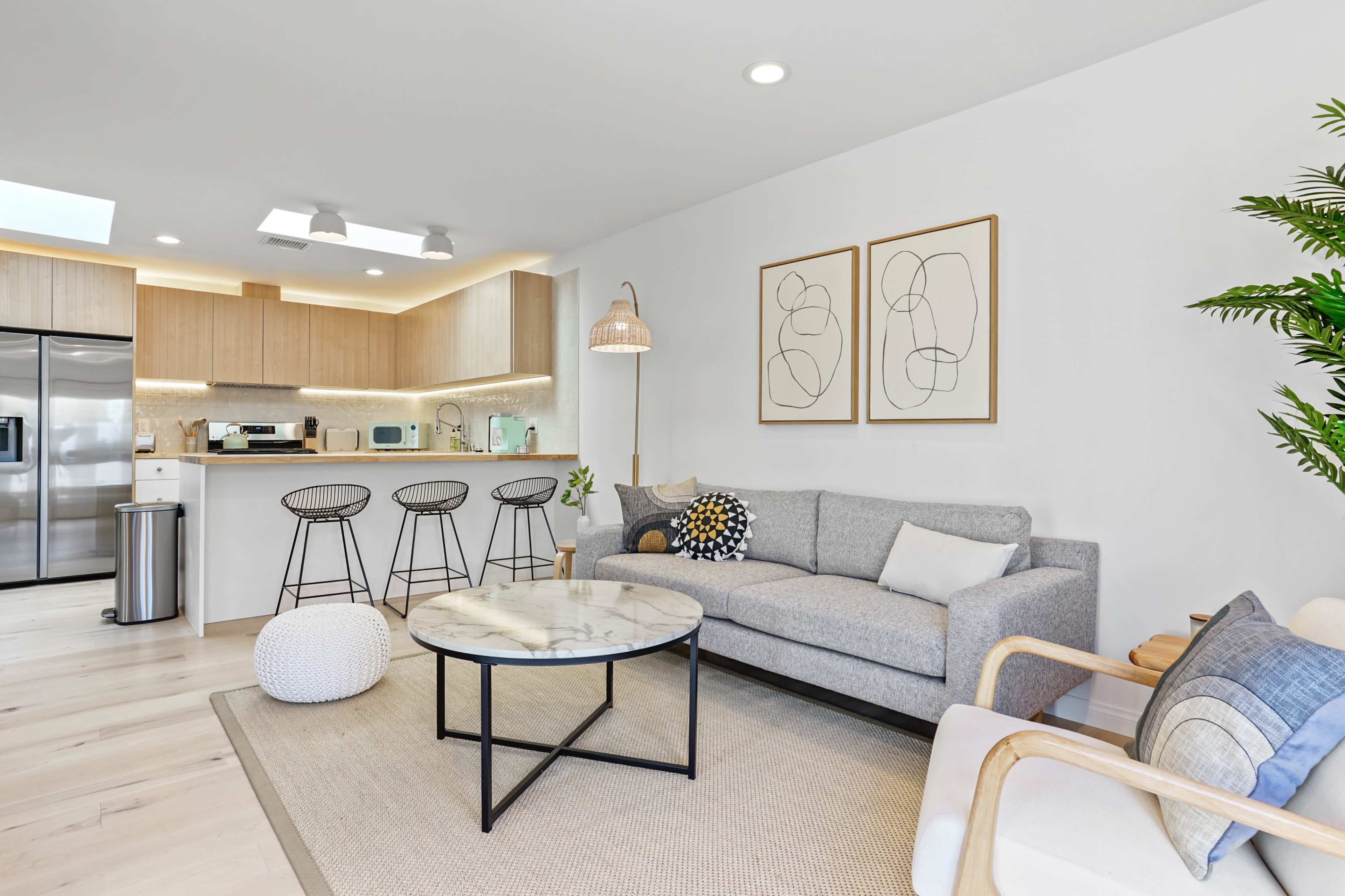 A modern living area featuring a gray sofa, a marble coffee table, two framed artworks on the wall, and a kitchen area with wooden cabinets.