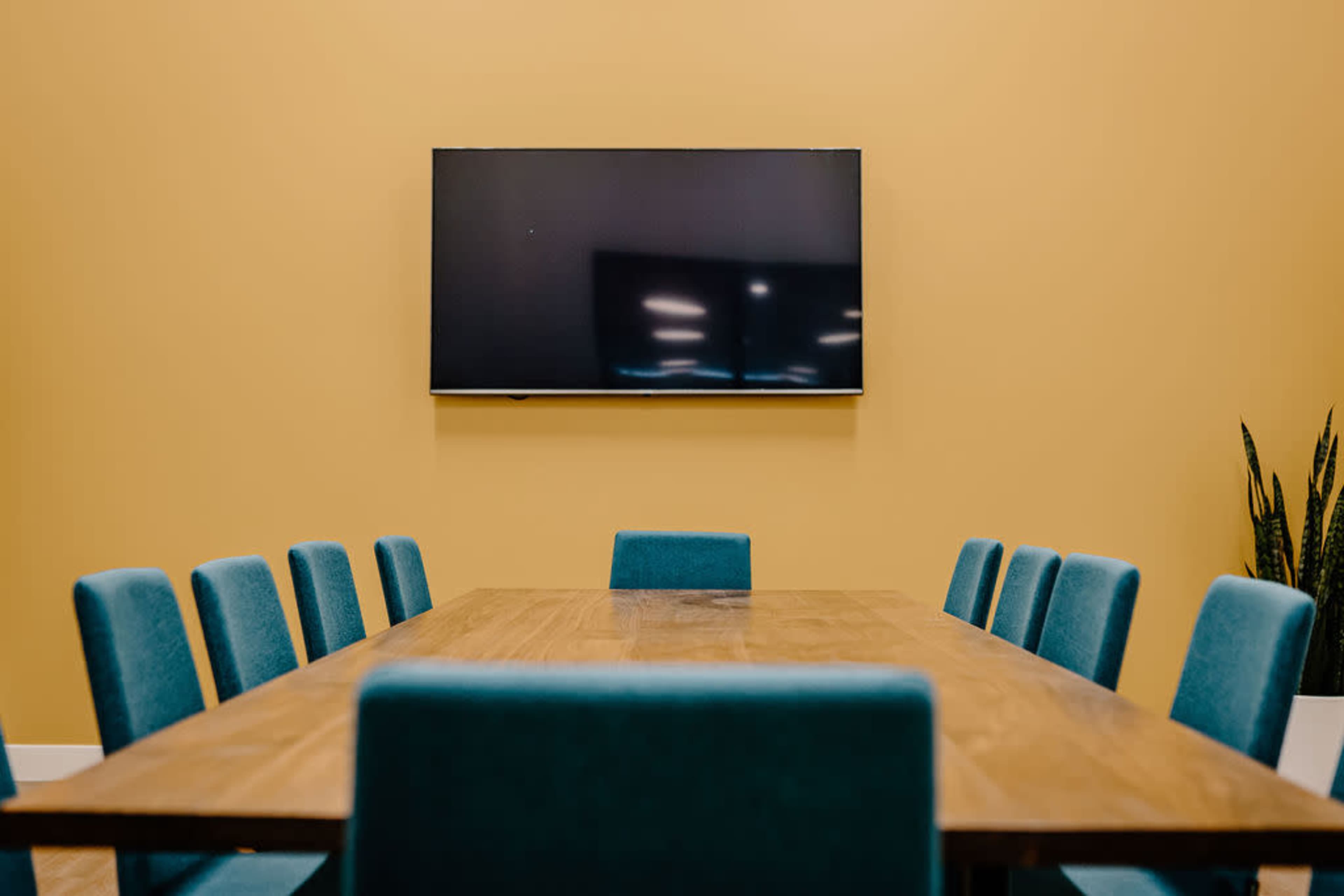 A wooden conference table is surrounded by teal chairs in a meeting room with a yellow wall and a flat-screen TV mounted above.