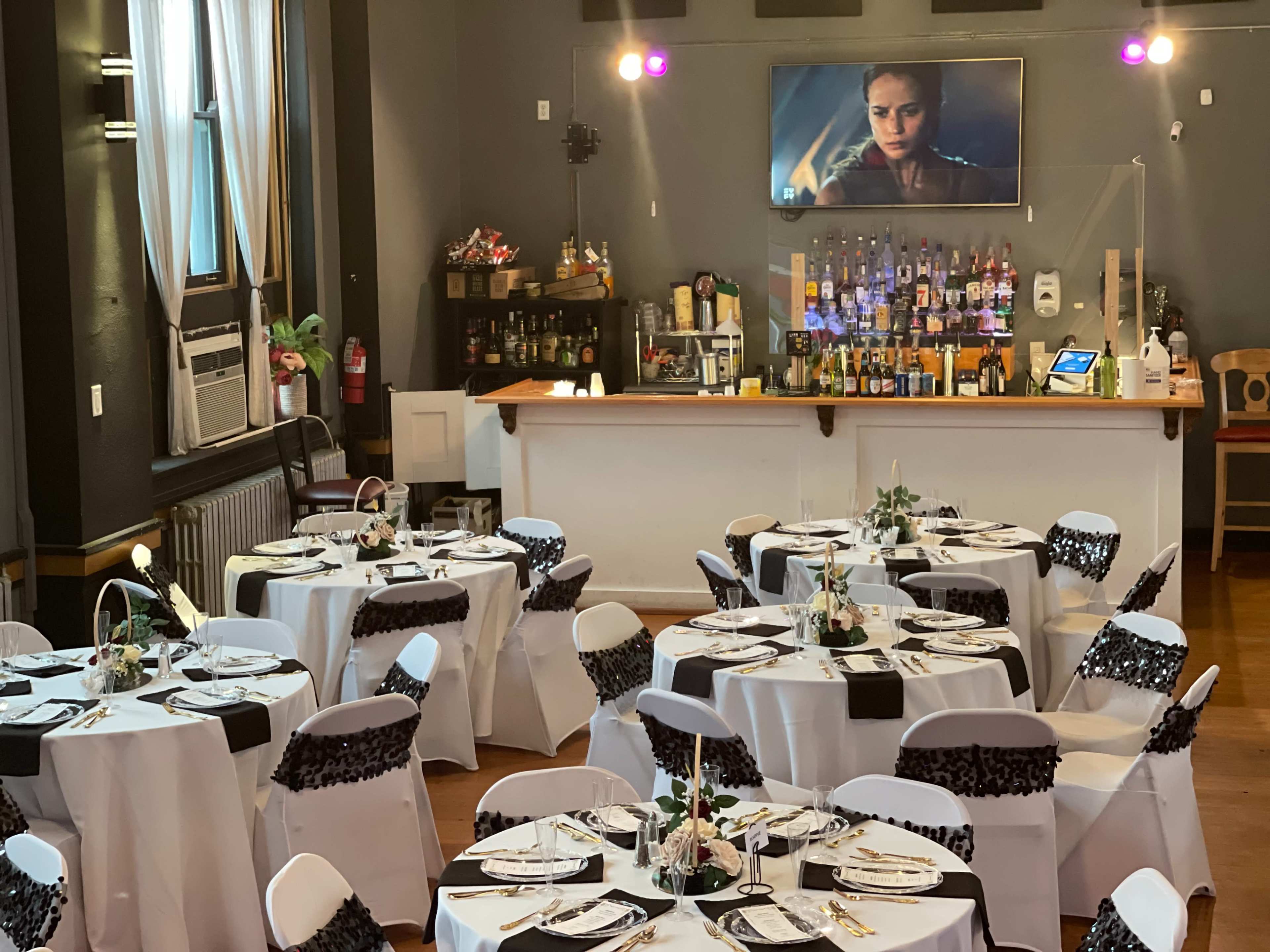 A dining area is set up with round tables covered in white tablecloths and black accents, with a bar and a screen displaying a scene in the background.