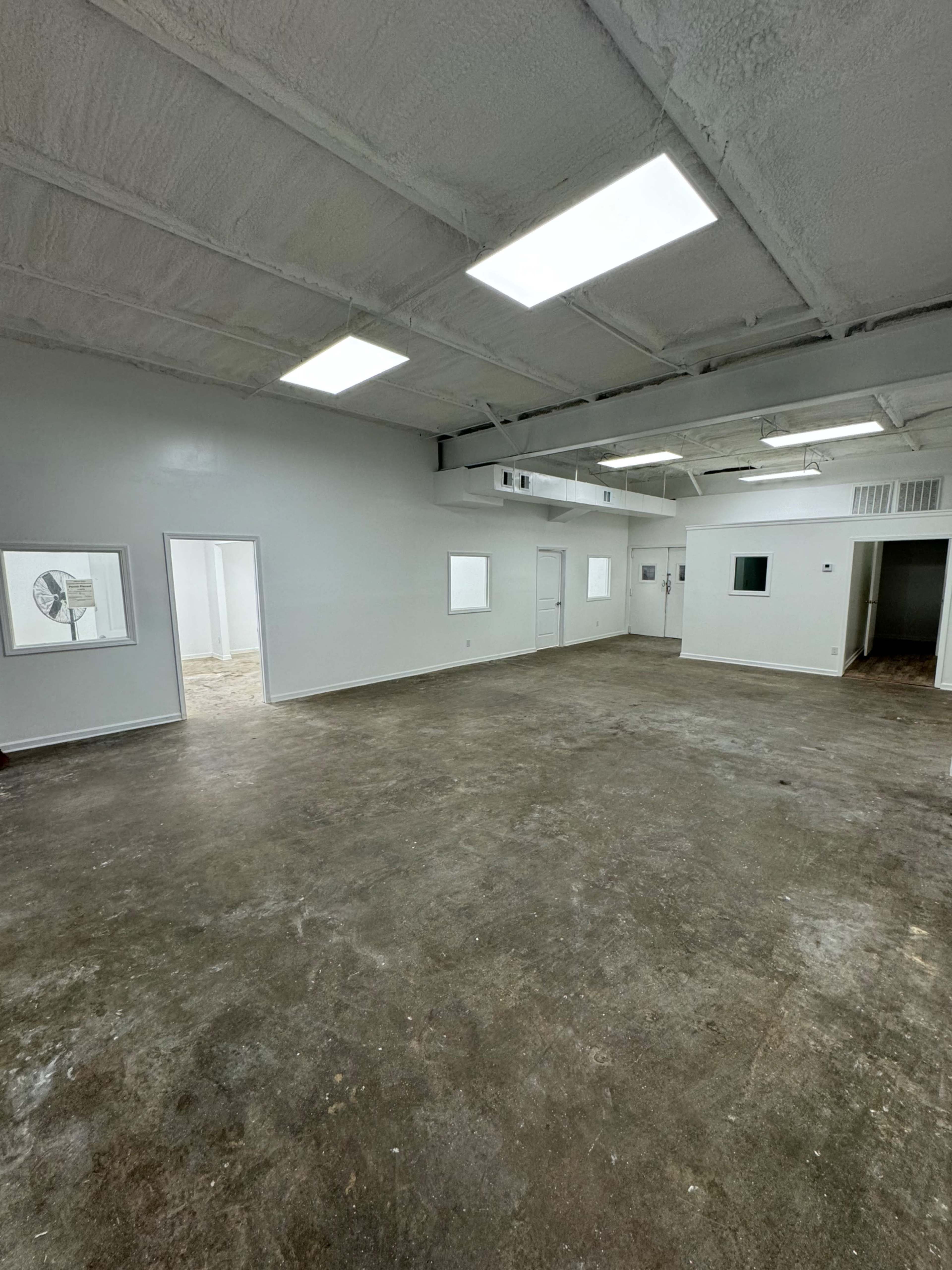 The image shows an empty, well-lit room with concrete flooring and white walls, featuring multiple doorways leading to other areas.