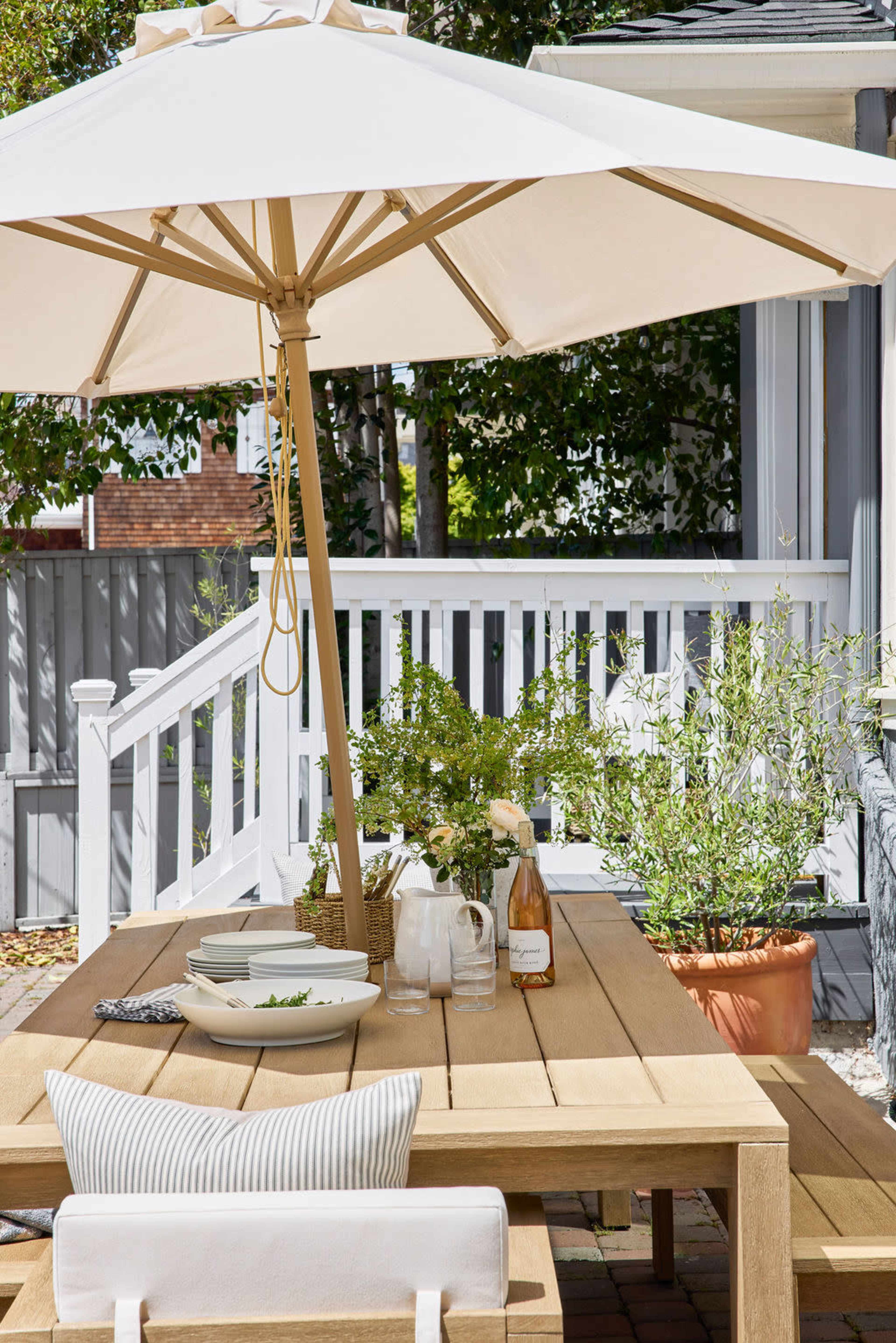 A wooden outdoor dining table is set with plates and drinks under a large umbrella, surrounded by greenery and a patio area.