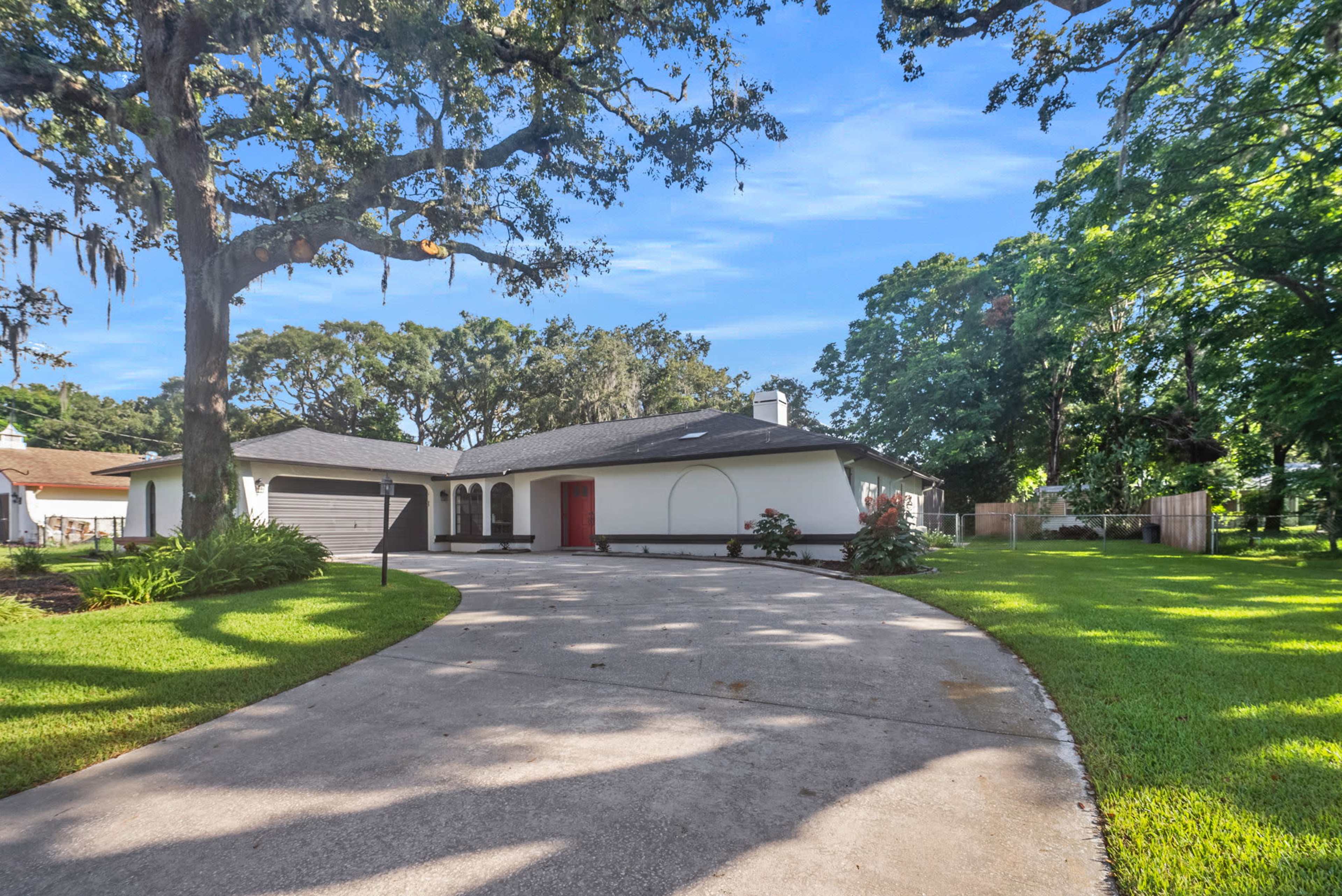 A single-story house with a red front door and a circular driveway is surrounded by green grass and large trees.
