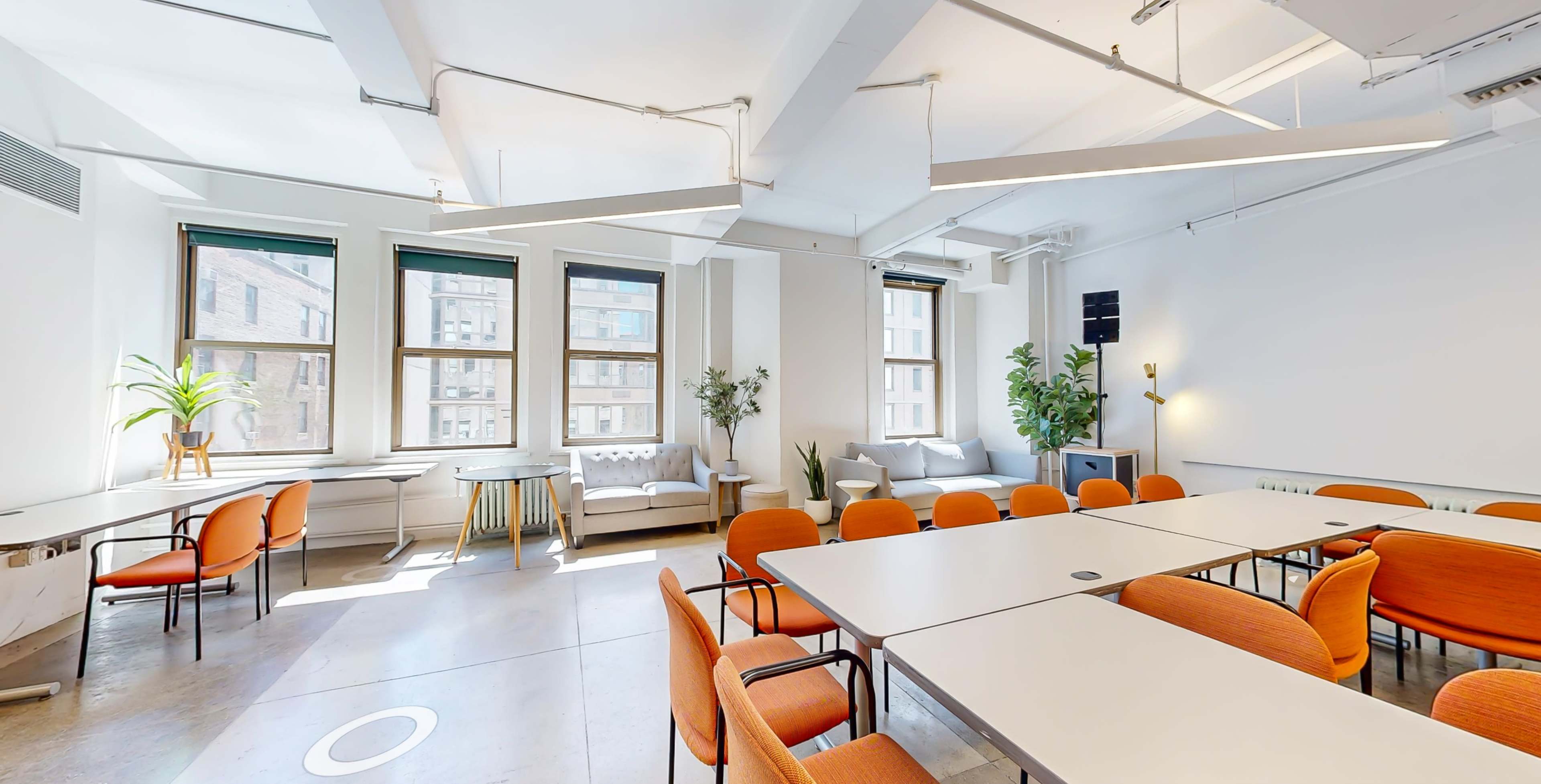 A bright meeting room with multiple tables and orange chairs arranged around them, large windows, and plants.