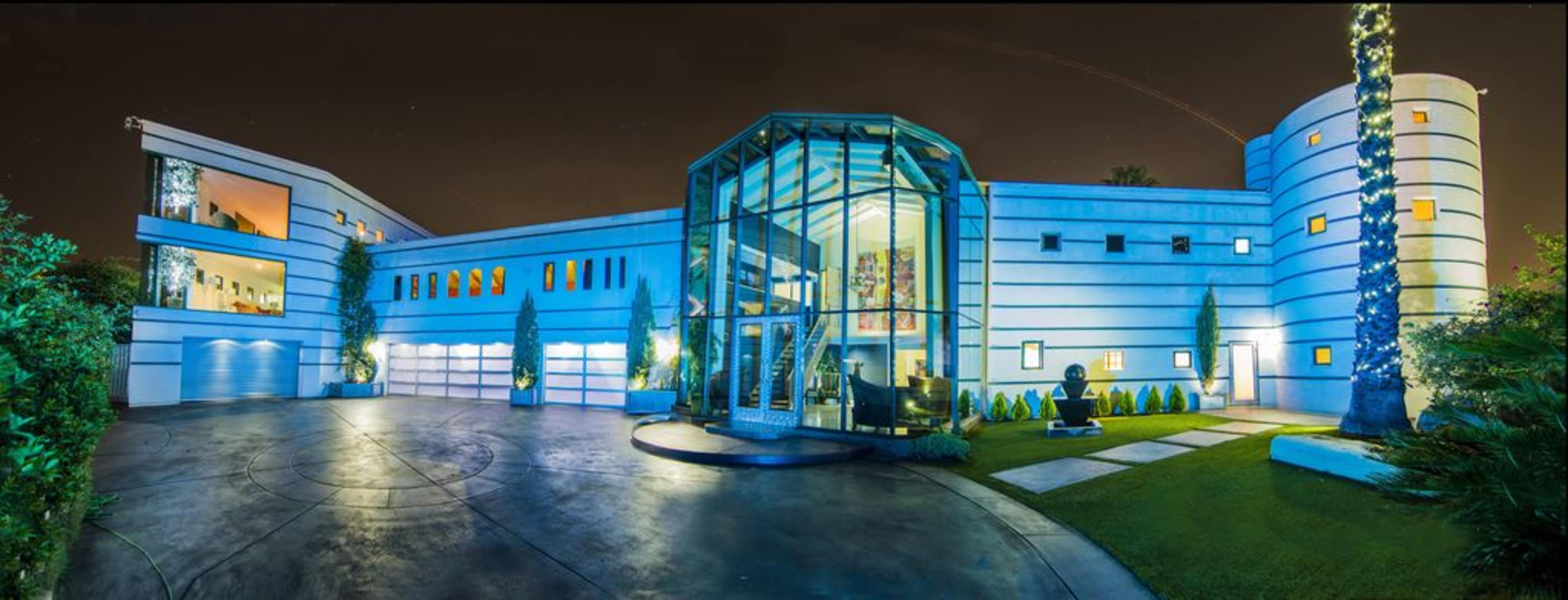 A modern residential building with a glass entrance, surrounded by landscaped gardens and palm trees, illuminated at night.