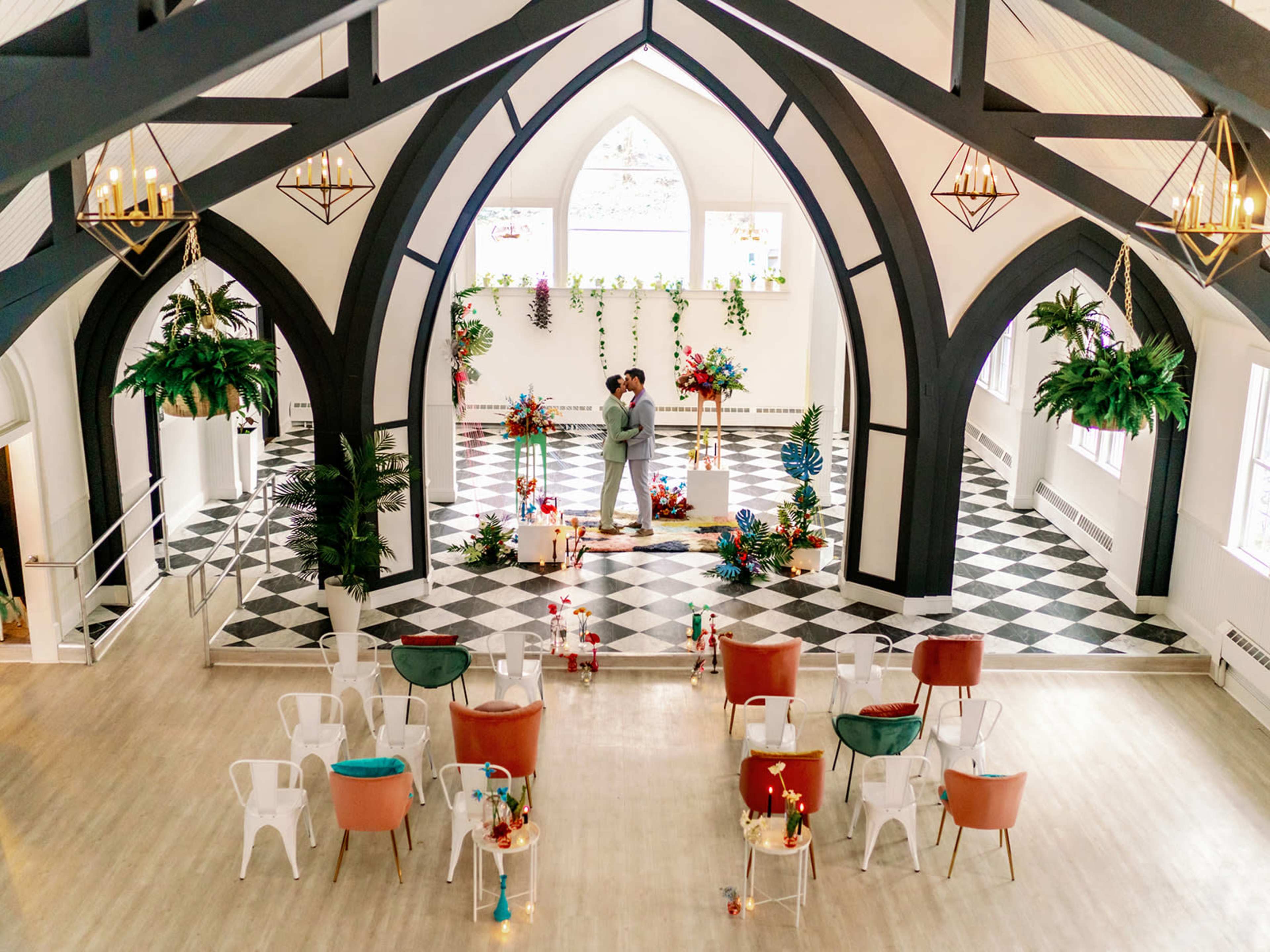 A couple embraces on a decorated stage in a spacious indoor venue with black and white checkered flooring and floral arrangements.