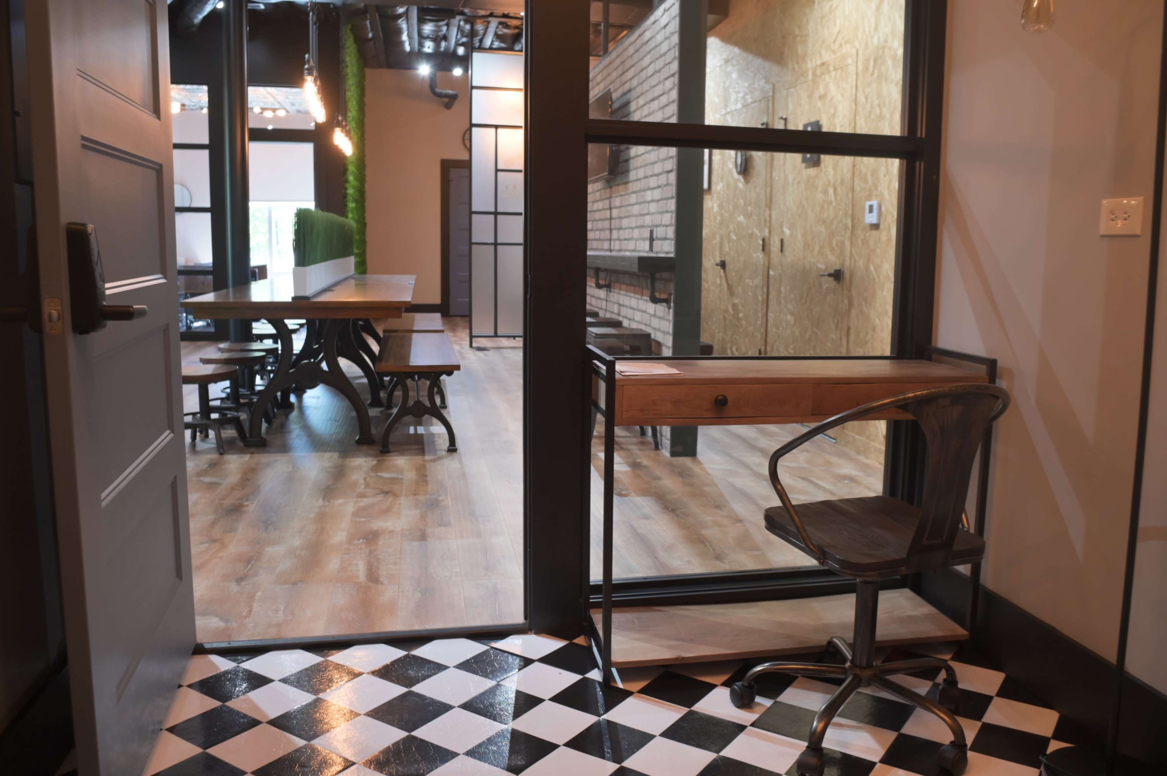 A modern workspace featuring a small desk with a rolling chair, viewed through a glass partition that opens to a large dining area with wooden tables in a contemporary setting.