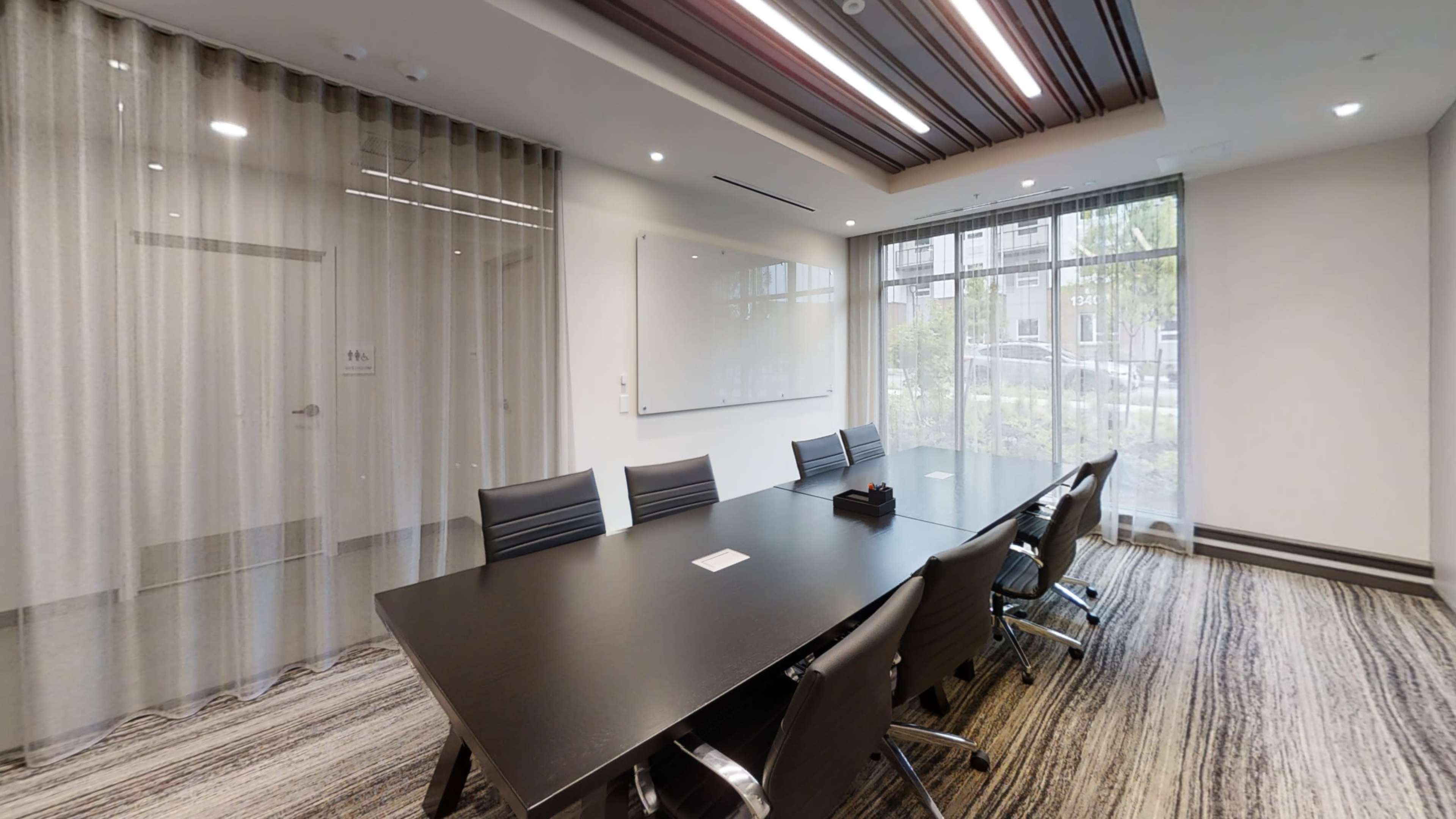 The image shows a modern conference room with a large black table surrounded by eight black chairs, and large windows allowing natural light to enter the space.